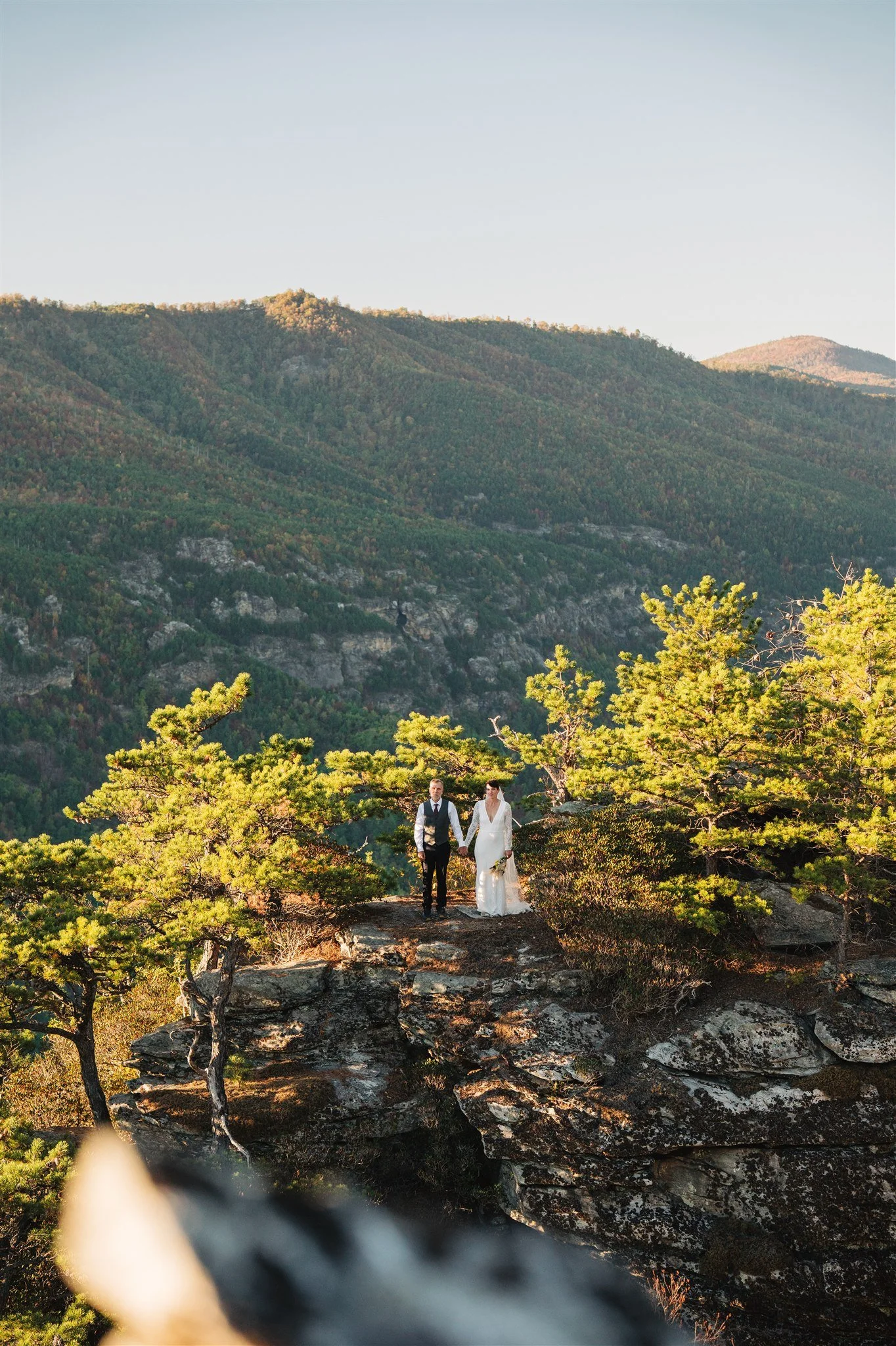 A bride and groom holding hands on a rocky ledge surrounded by green trees with a mountain landscape in the background during sunset.