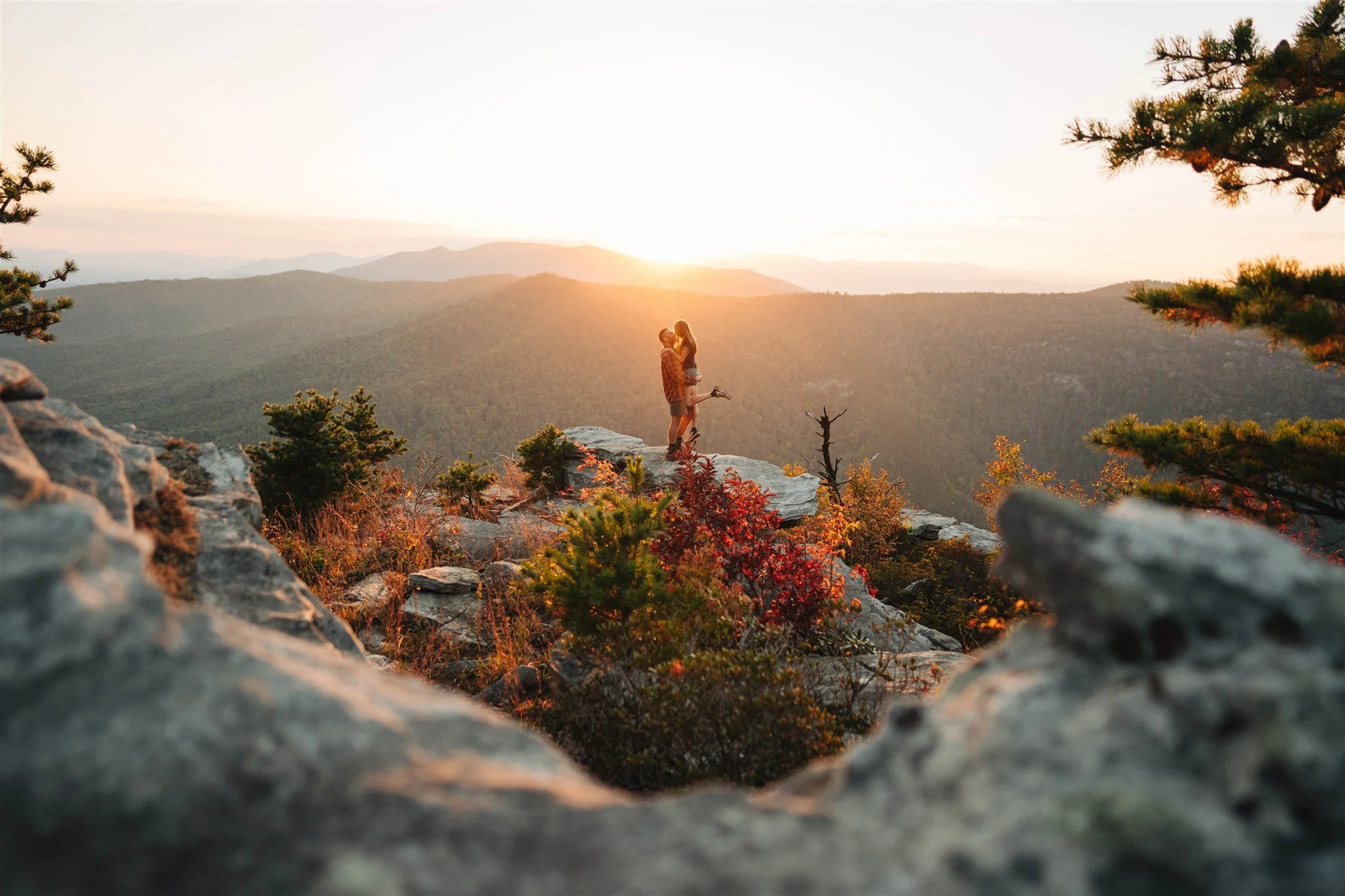 A couple standing on a rocky ledge overlooking an expansive mountain landscape at sunset, surrounded by trees with autumn foliage.