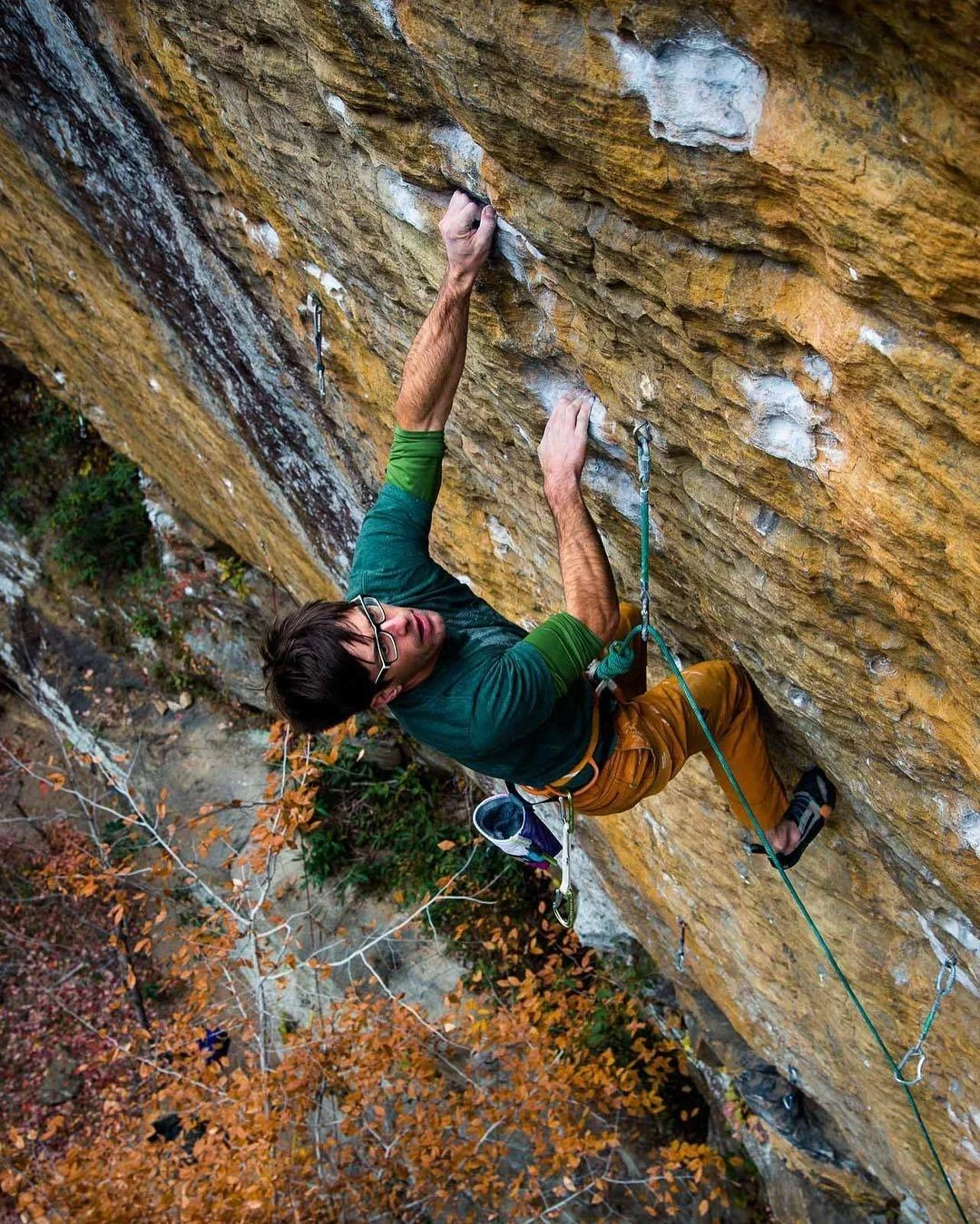 Person rock climbing on a steep cliff with safety ropes, surrounded by autumn foliage.