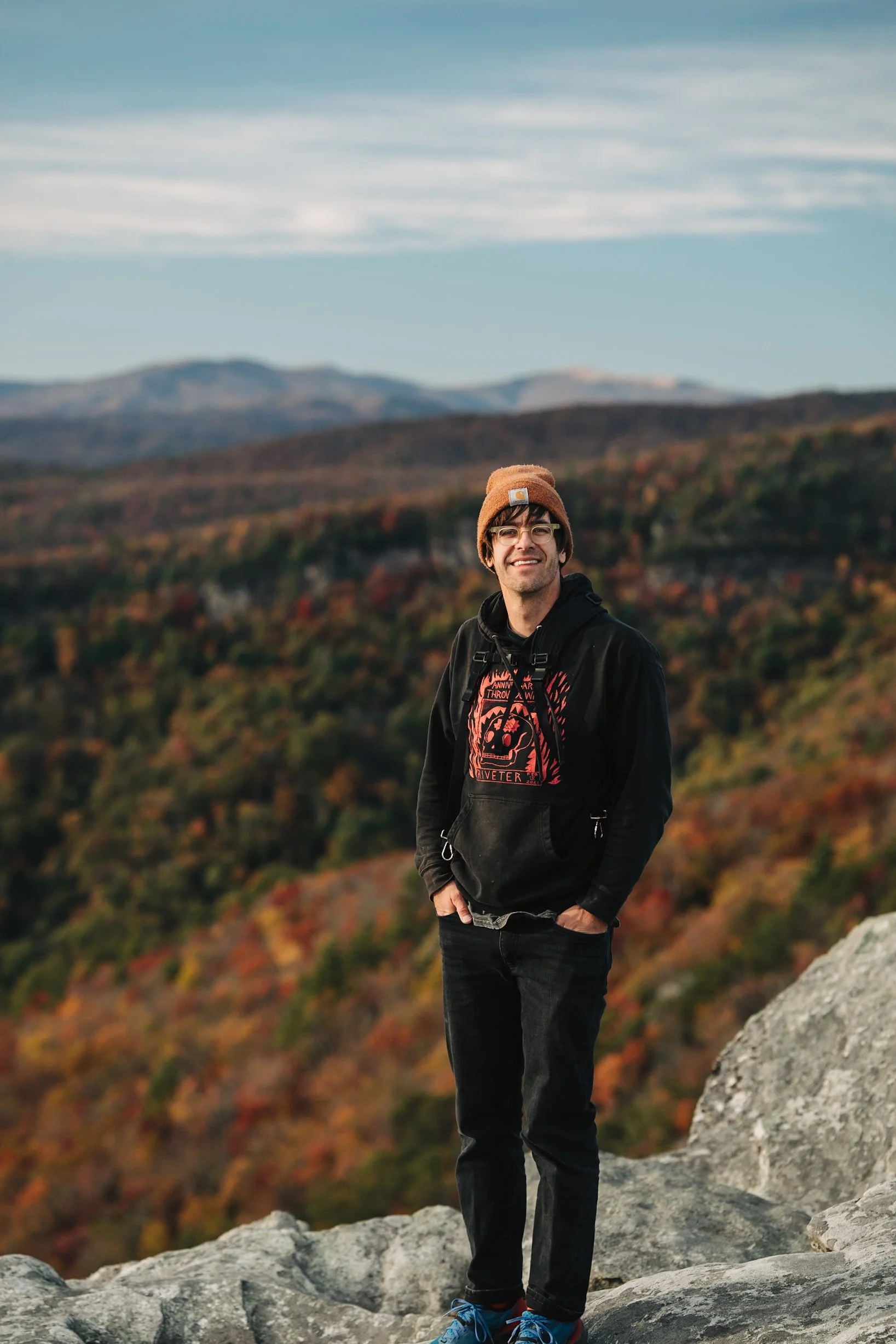 A smiling man standing on a rocky ledge in front of autumn-colored mountains under a cloudy sky.
