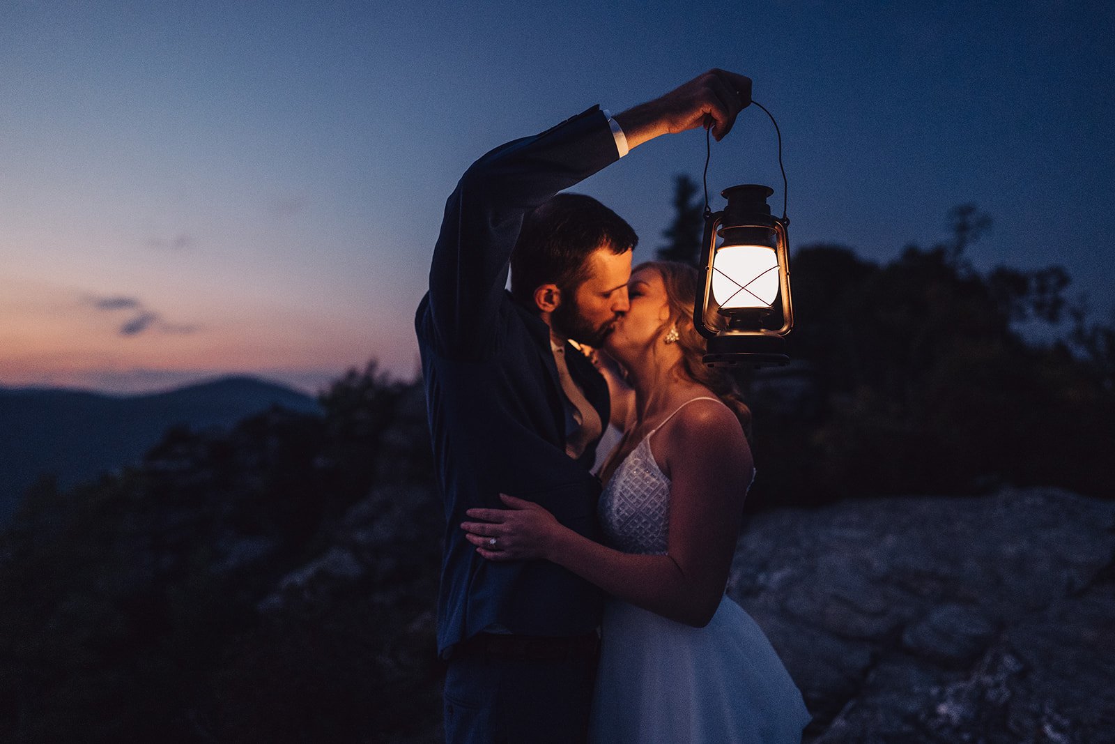 A couple sharing a kiss at dusk, holding a lantern, with a scenic mountainous background.