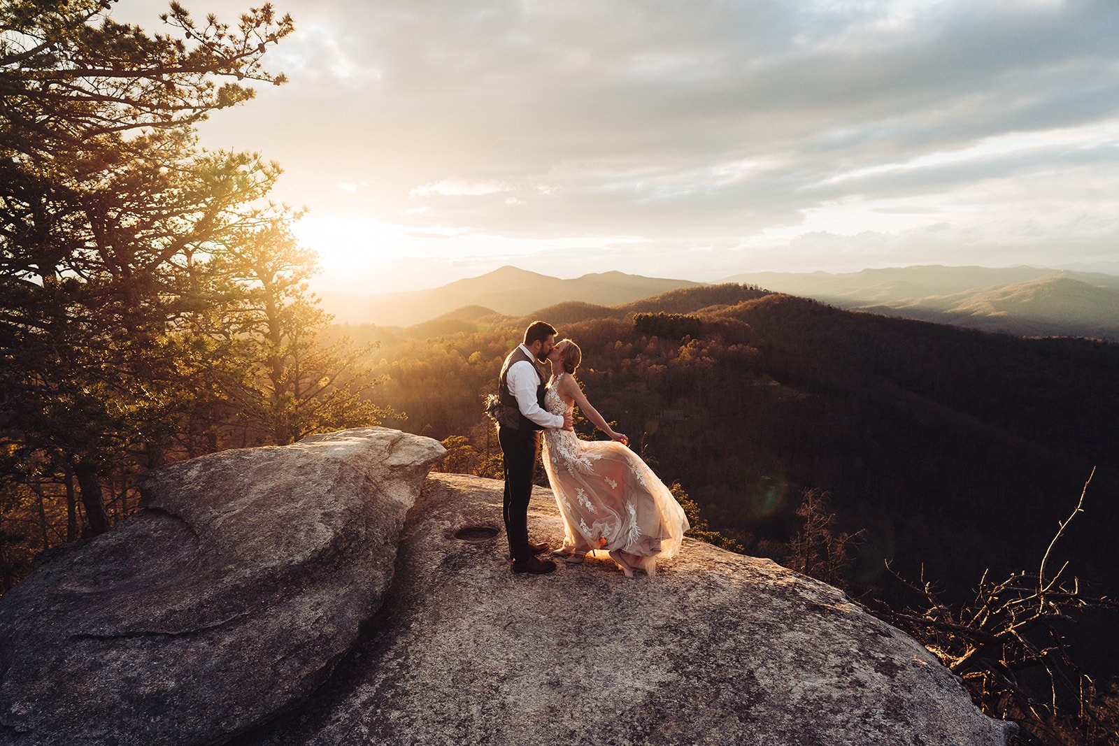 A couple dressed in wedding attire standing on a large rock at sunset with a mountain landscape in the background.