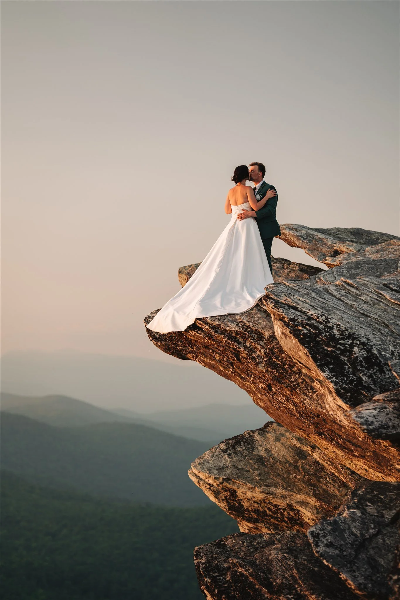 A bride and groom kiss on a rocky cliff at sunset, overlooking a mountain landscape.