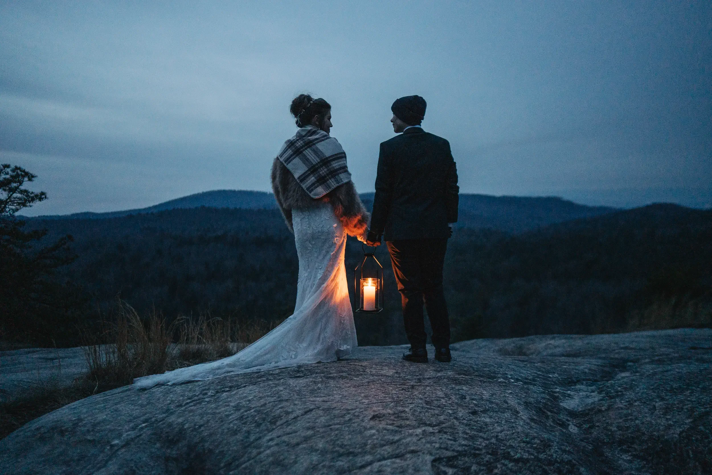 A couple is standing outdoors at dusk, holding hands and gazing at each other. The woman is wearing a white dress and a plaid shawl, while the man is dressed in a black coat and beanie. They are holding a lantern with a lit candle.