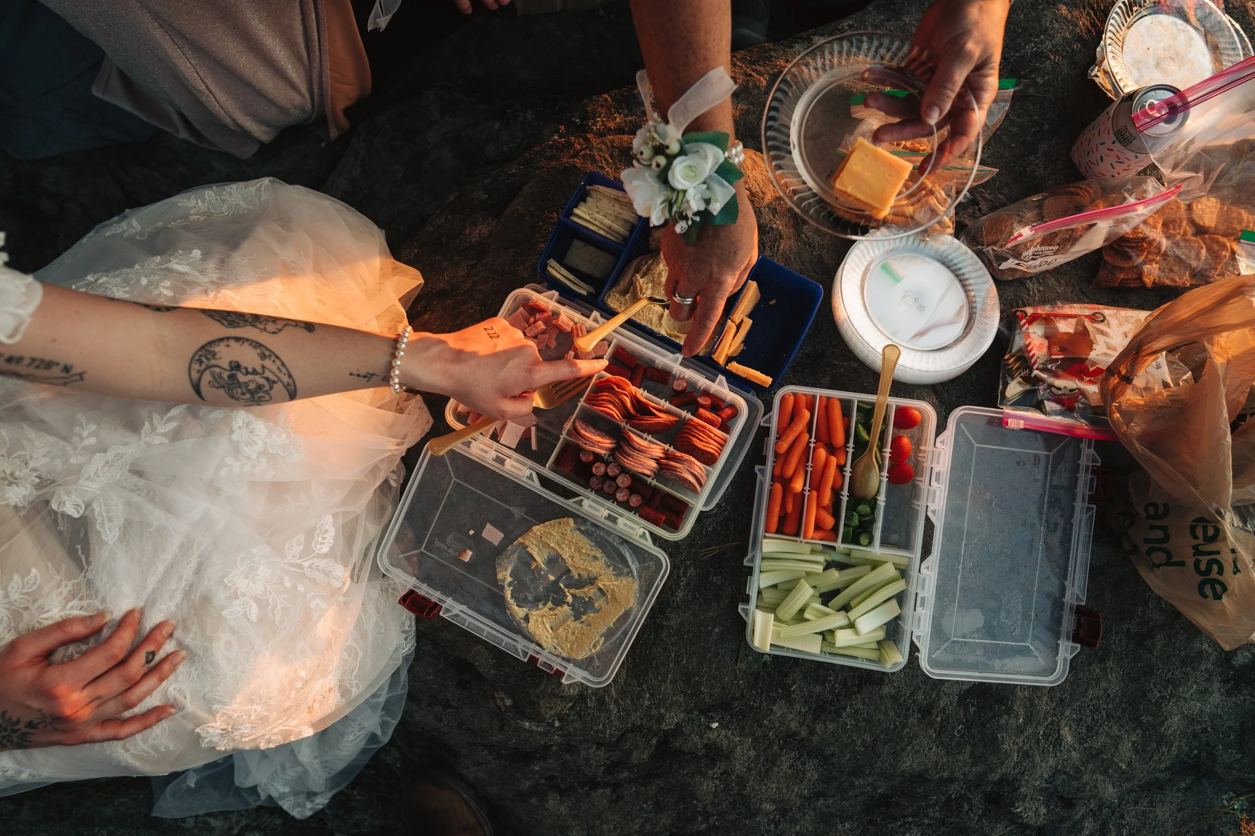 Top-down view of a picnic setup on a dark surface with food containers, snacks, and drinks, including vegetables, sliced meats, cheese, and a candle, with two people's hands reaching for food.