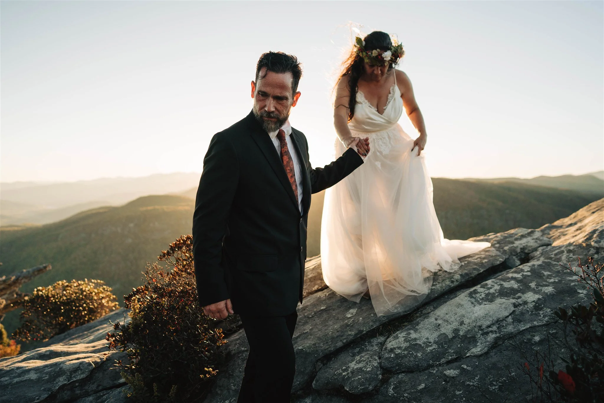 A groom holding a bride's hand as she steps down from rocks on a mountain at sunset. The bride is wearing a white wedding dress and a floral crown. The scene features a scenic mountain landscape in the background.