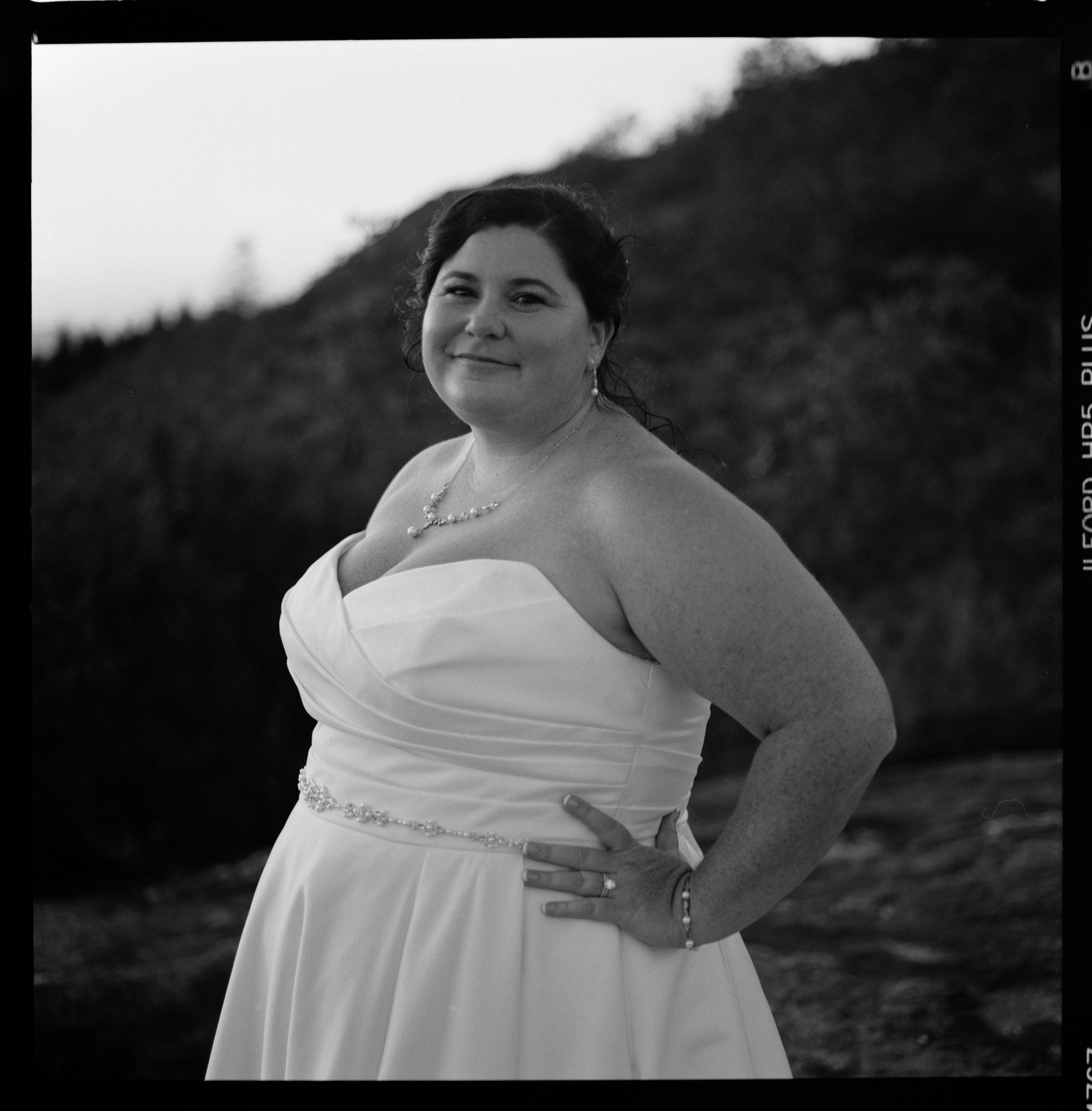 A smiling woman in a strapless wedding dress standing outdoors with a hill in the background, wearing jewelry including earrings, a necklace, bracelet, and ring.