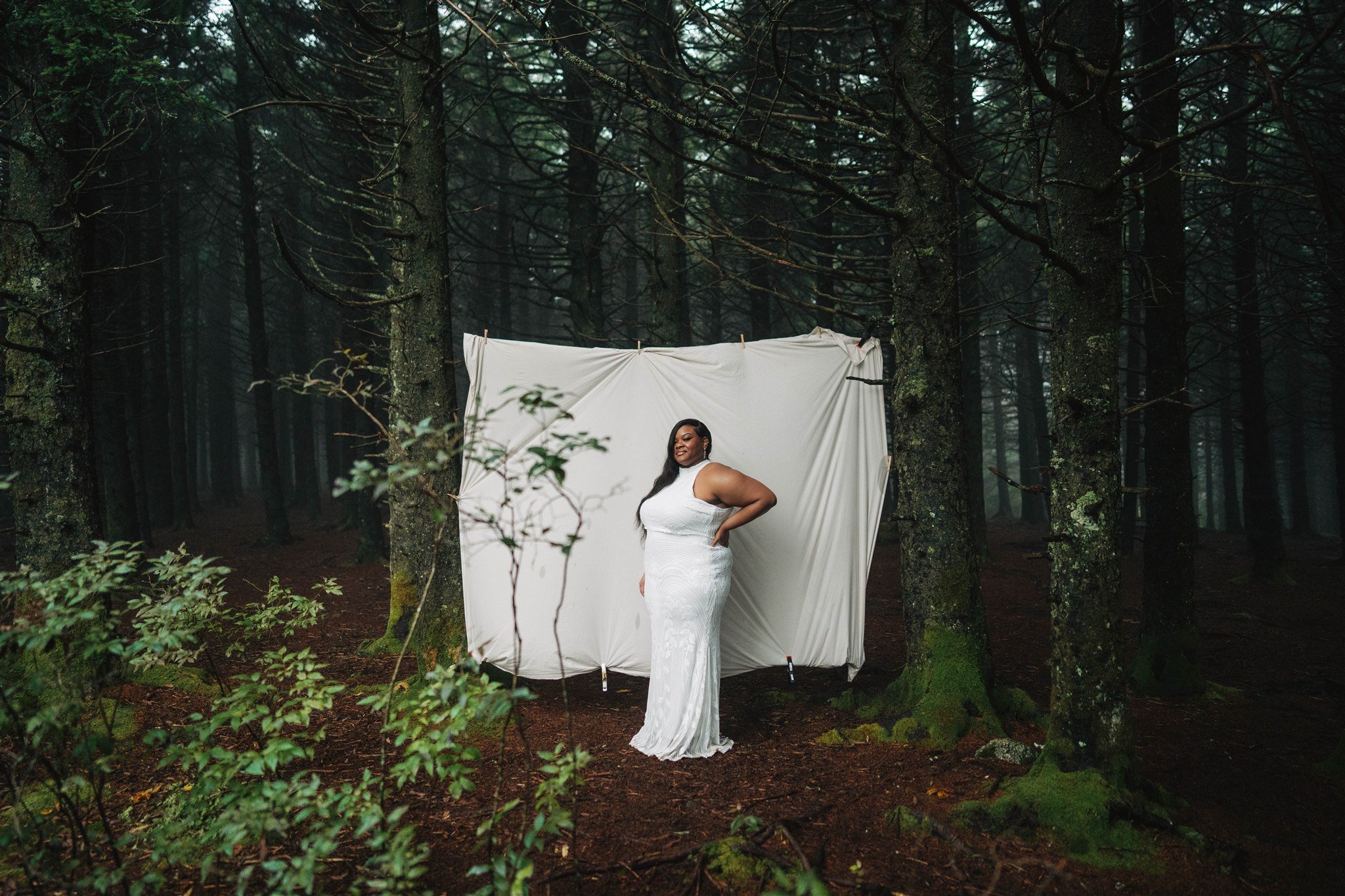 Woman in a white gown standing in front of a white cloth backdrop in a forest.