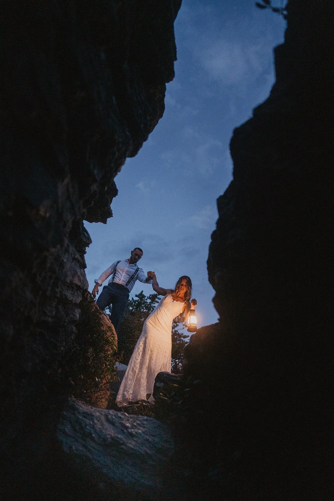 A couple, dressed in wedding attire, holding hands and standing outdoors during twilight, with the woman holding a lantern, framed by large rocks creating a natural opening.