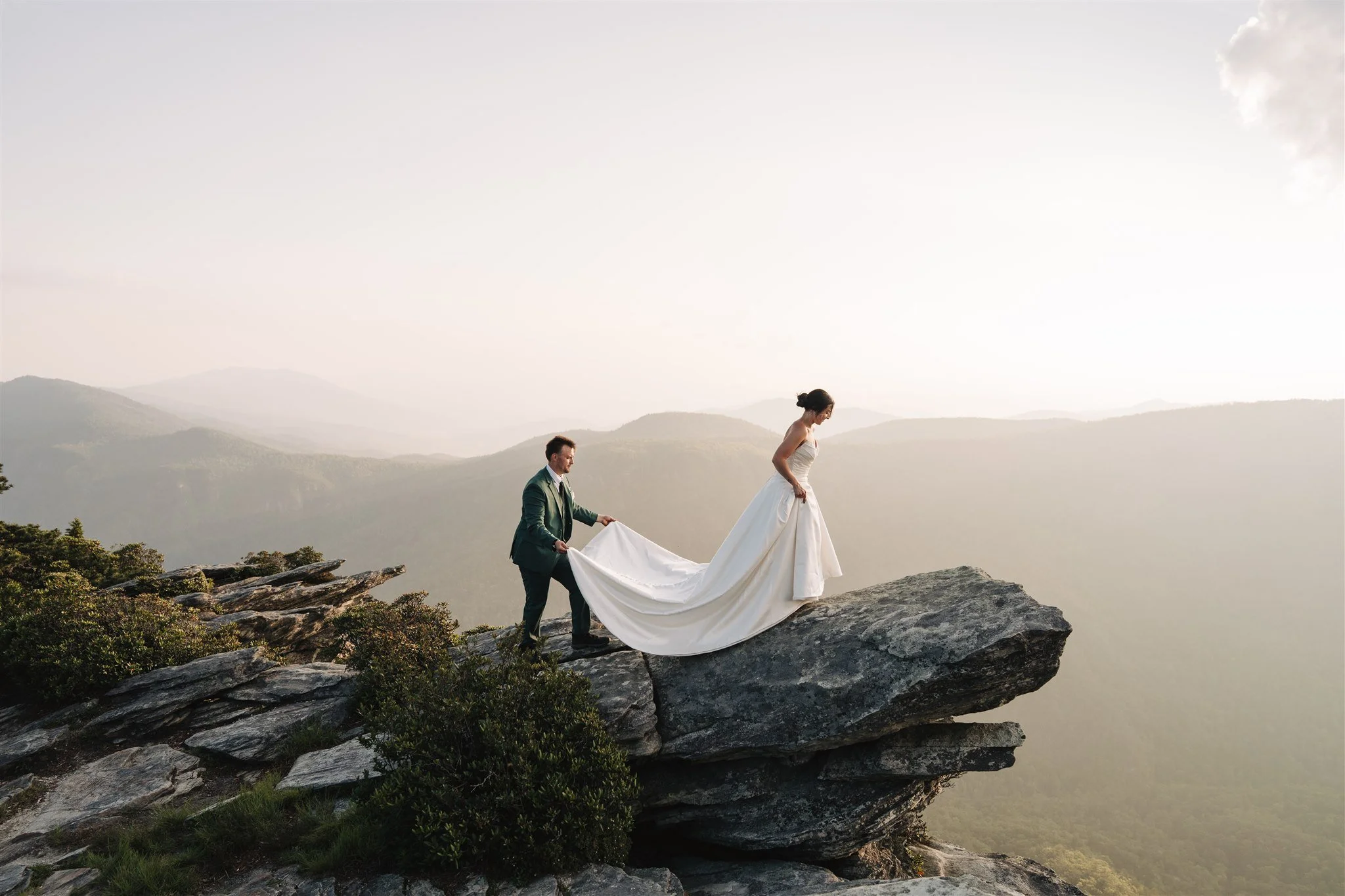 A bride and groom standing on a large rock overlooking a mountain landscape. The groom is adjusting the bride's wedding dress as she stands on the edge of the rock. The scene is set during daylight with a hazy sky.