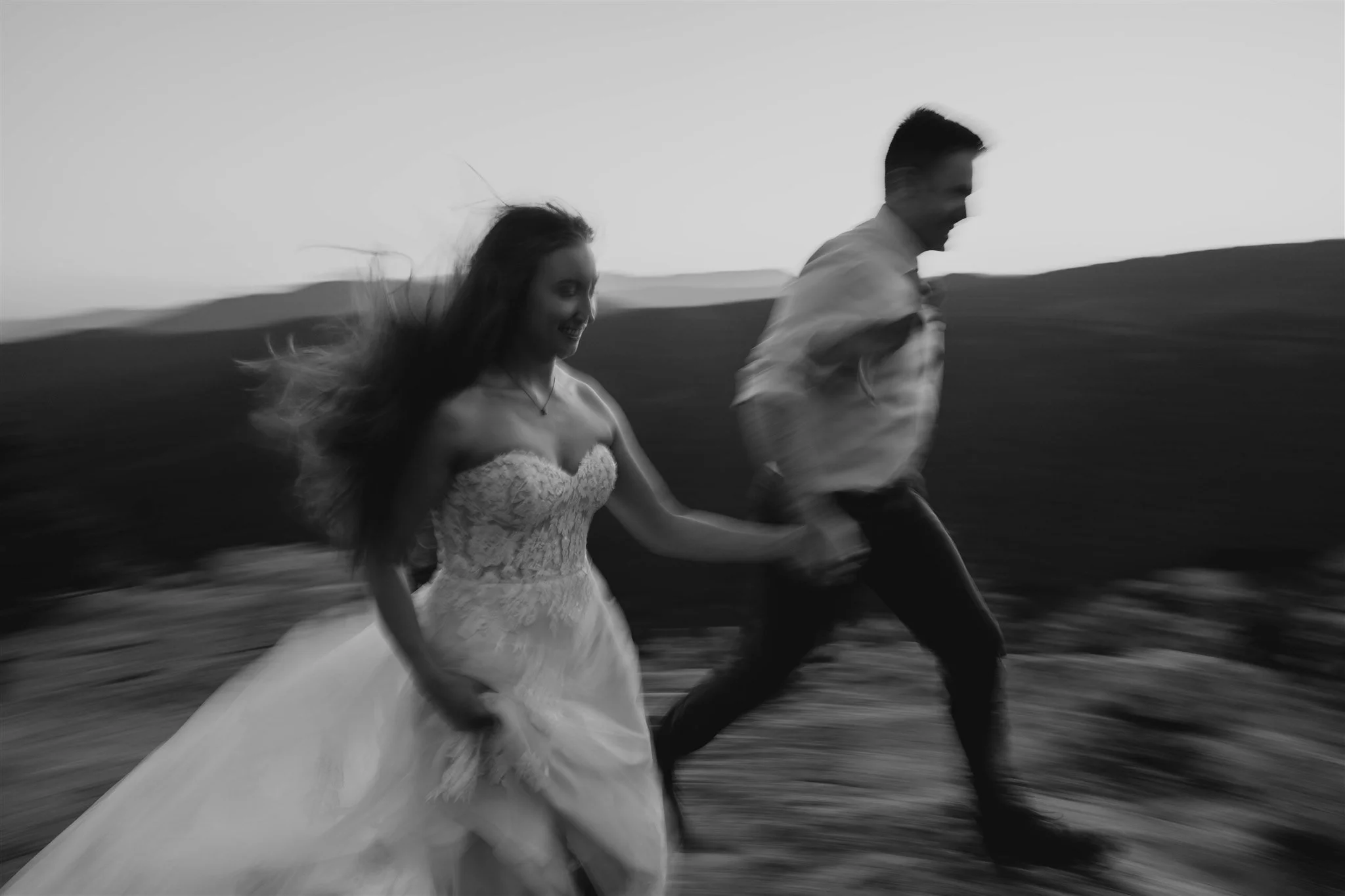 Blurred black and white photo of a couple running, the woman wears a wedding dress, and the man is in formal attire, with mountains in the background.