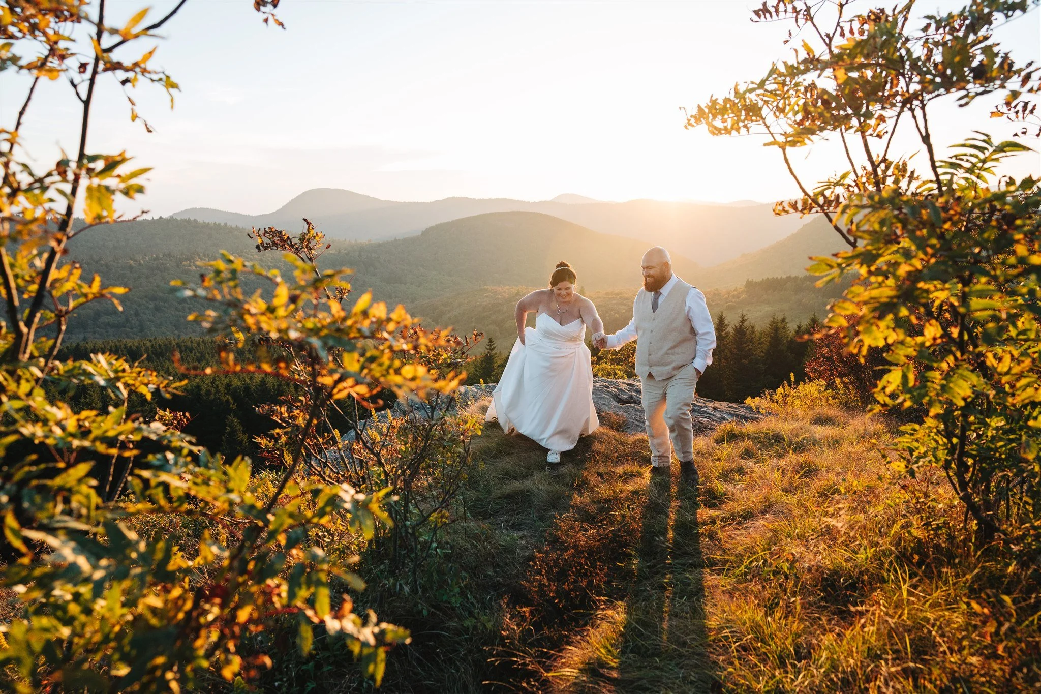 A bride and groom walking hand in hand on a grassy hillside during sunset surrounded by mountains and trees.
