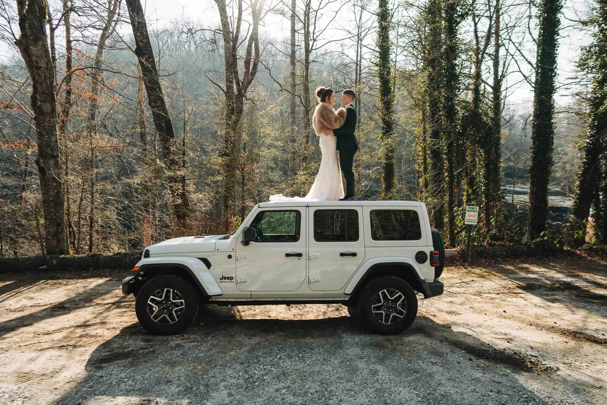 A bride and groom standing on top of a white Jeep in a wooded area, with the bride in a wedding dress and fur stole, and the groom in a suit, facing each other lovingly.