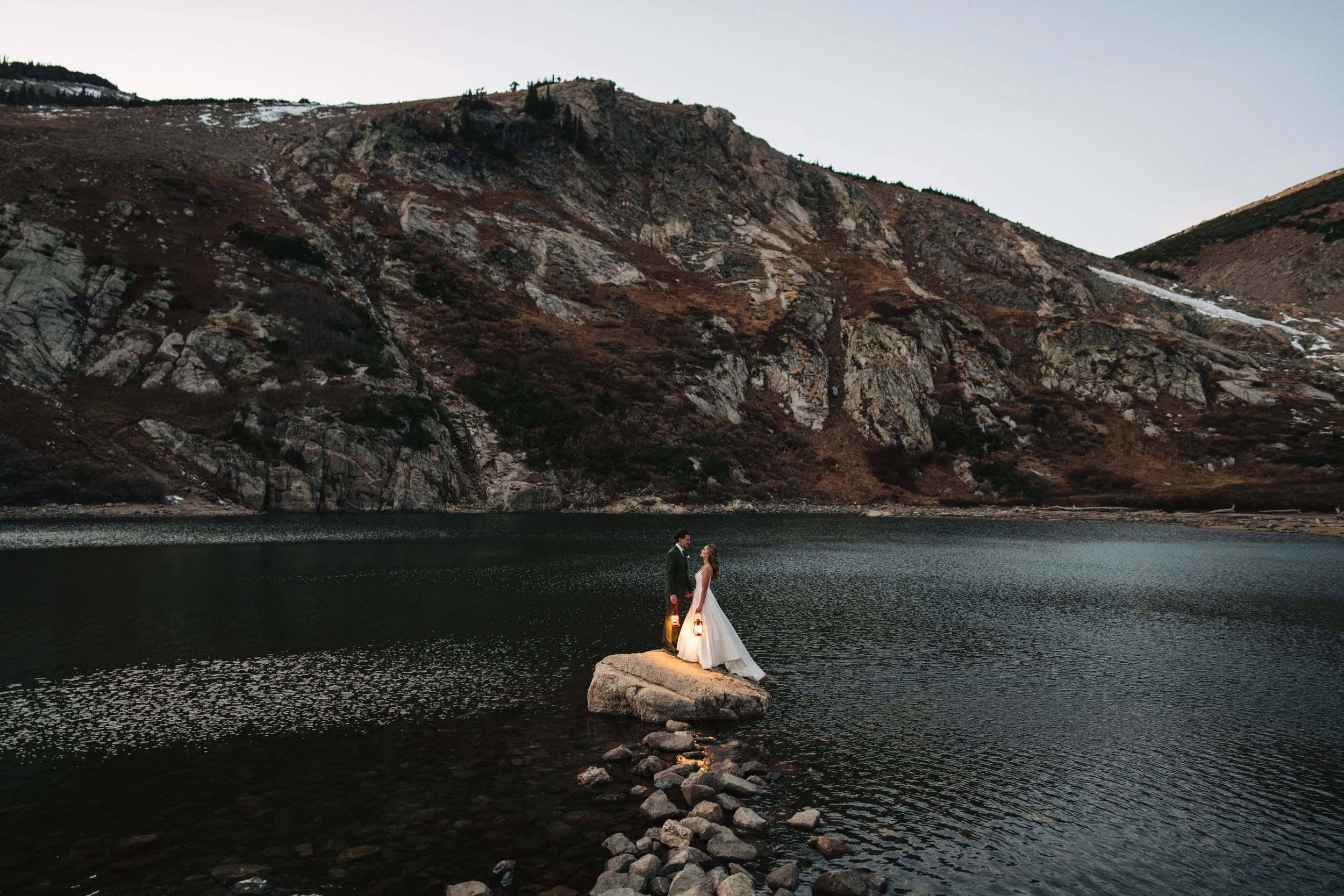A couple in wedding attire standing on a large rock in a lake, overlooking mountains at dusk.