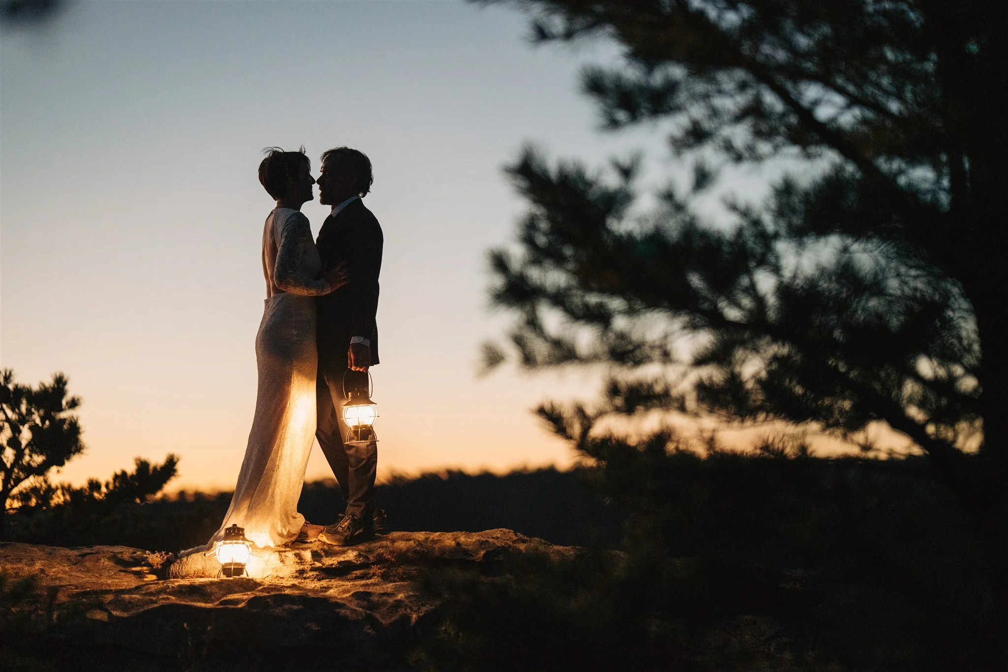 Silhouette of a couple in wedding attire standing close together on a rocky surface at sunset, holding lanterns, with trees in the background.