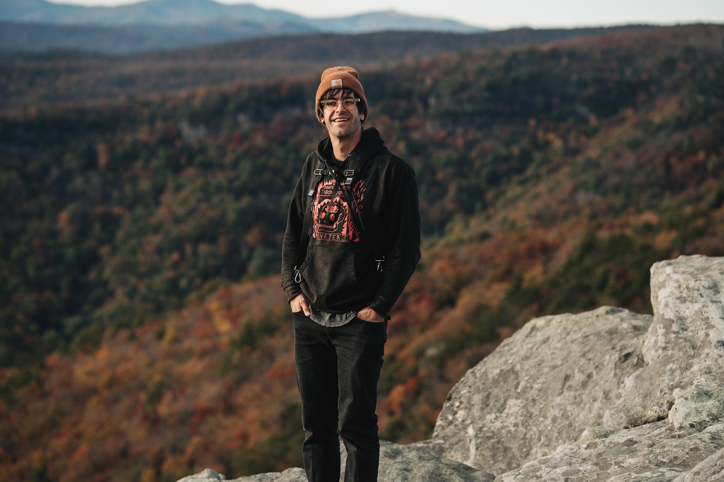 MountainWed owner and photographer, Andy Wickstrom, standing on a rocky overlook in a mountainous area with autumn foliage, wearing a tan beanie, glasses, a black hoodie with a graphic design, and black jeans, smiling at the camera.