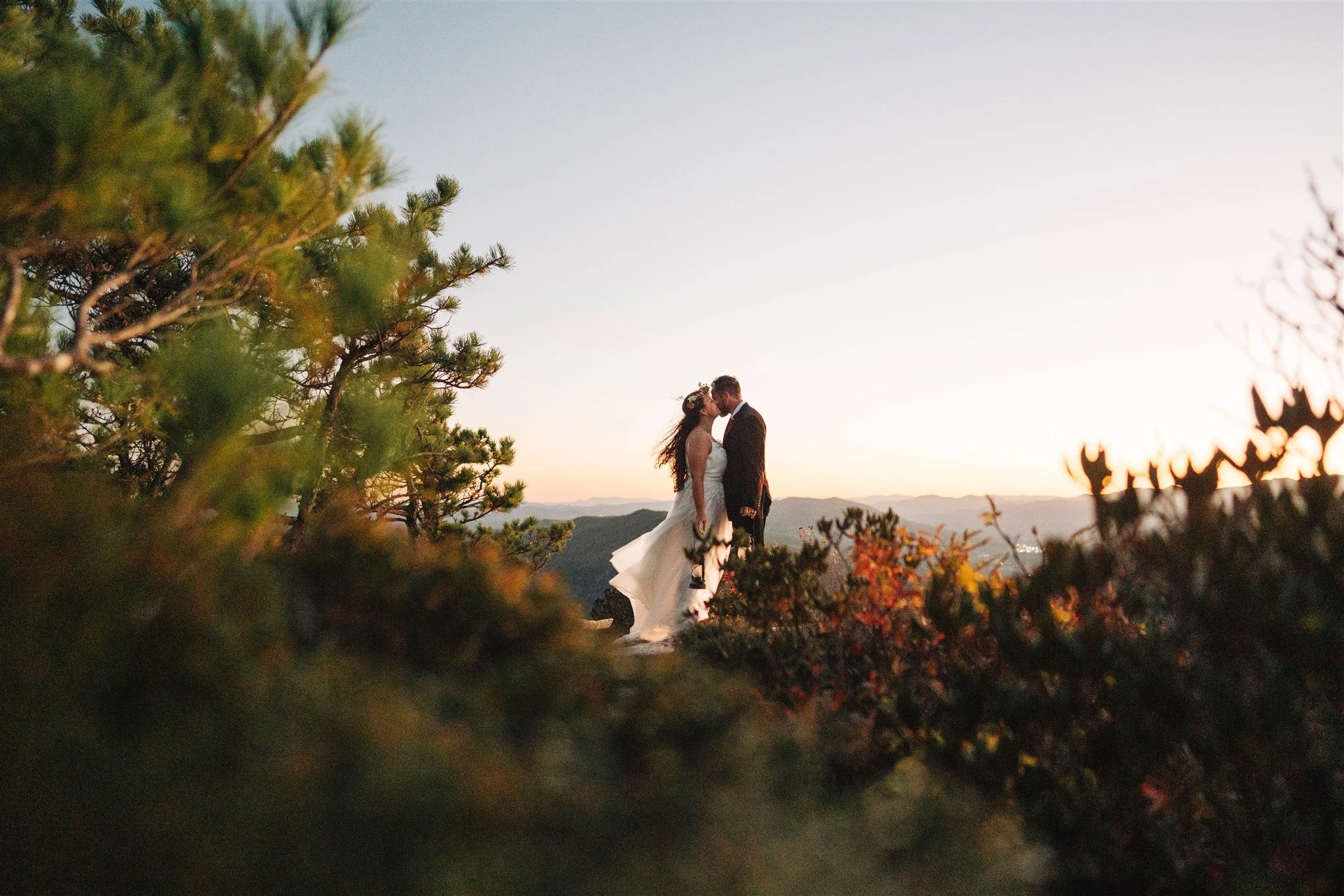 A bride and groom kissing in a scenic outdoor setting during sunset, framed by trees and bushes, with mountains in the background.