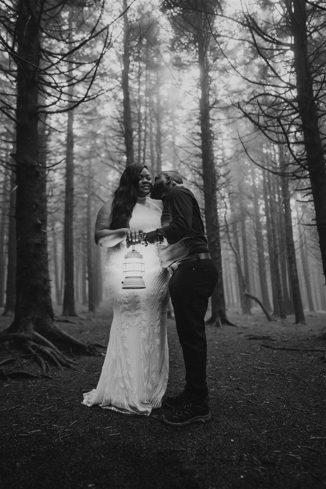 A black-and-white photo of a couple standing in a dense forest, with the woman holding a lantern. The woman is wearing a long, elegant dress, and the man is dressed casually. They are close together, sharing an intimate moment.
