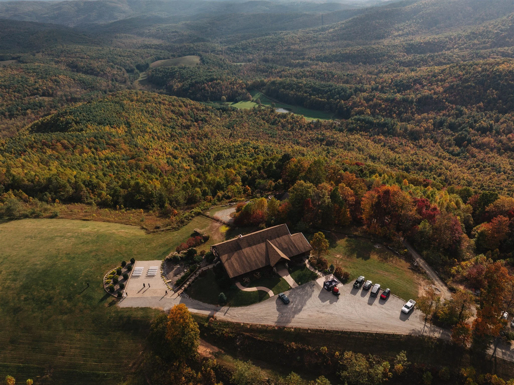 Aerial view of a rustic pavilion near a parking lot surrounded by colorful fall foliage in a mountainous landscape.