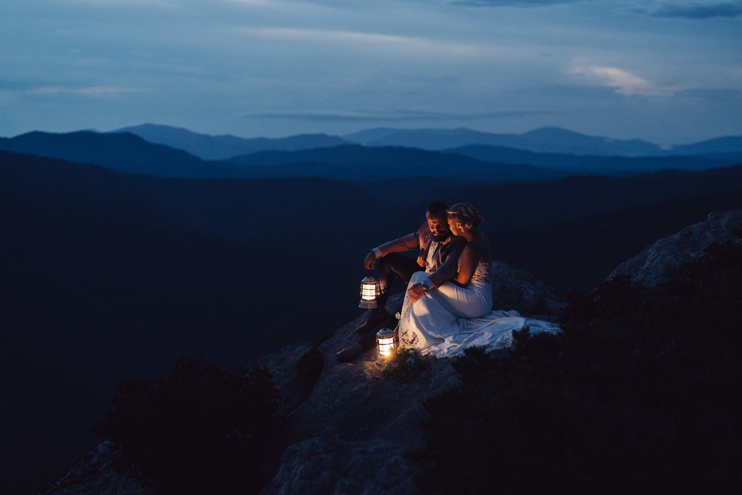 Newly married couple sitting together by lantern light on a mountain overlook after sunset