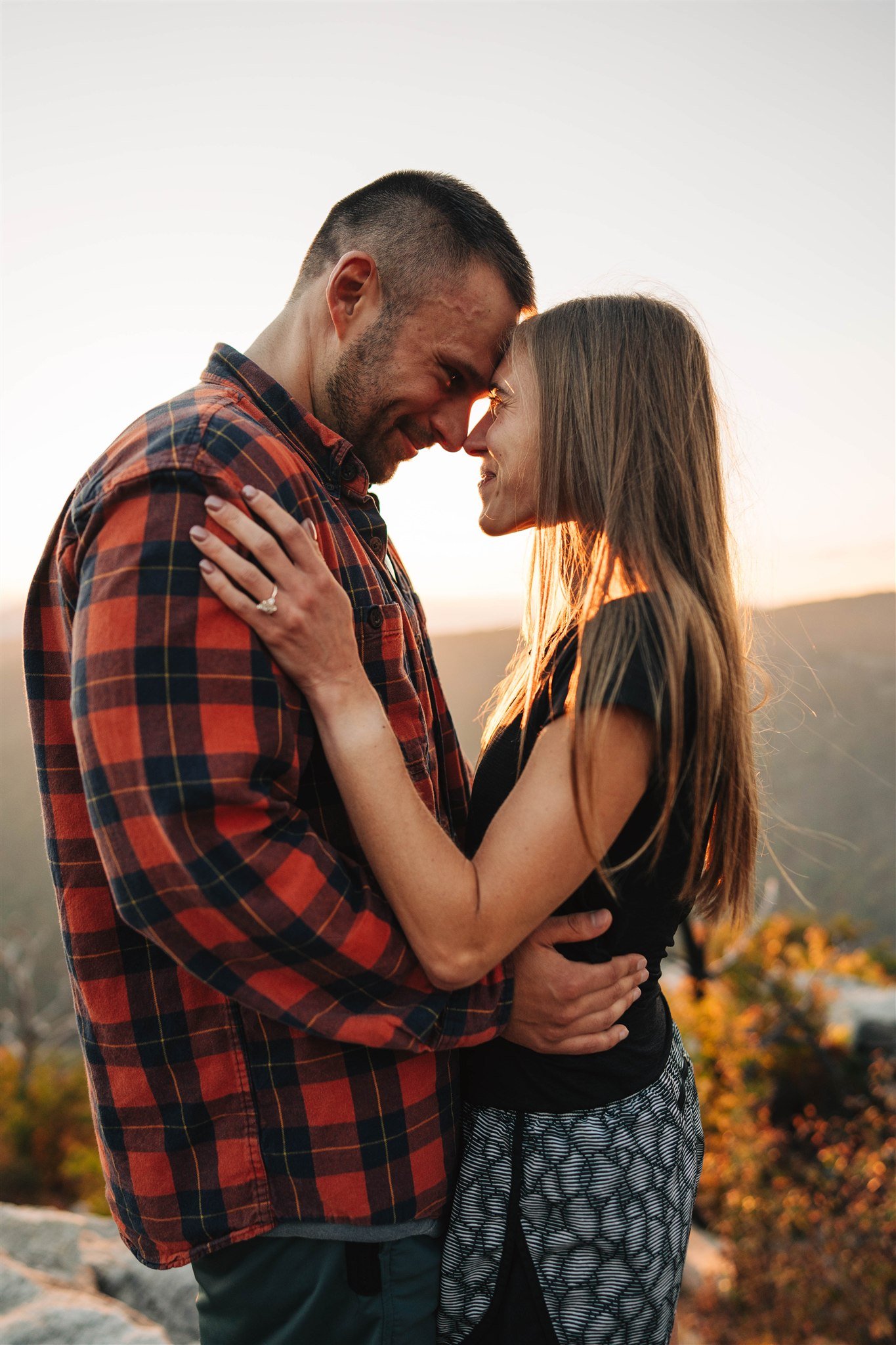 A couple standing close together with foreheads and noses touching, smiling at each other outdoors during sunset.
