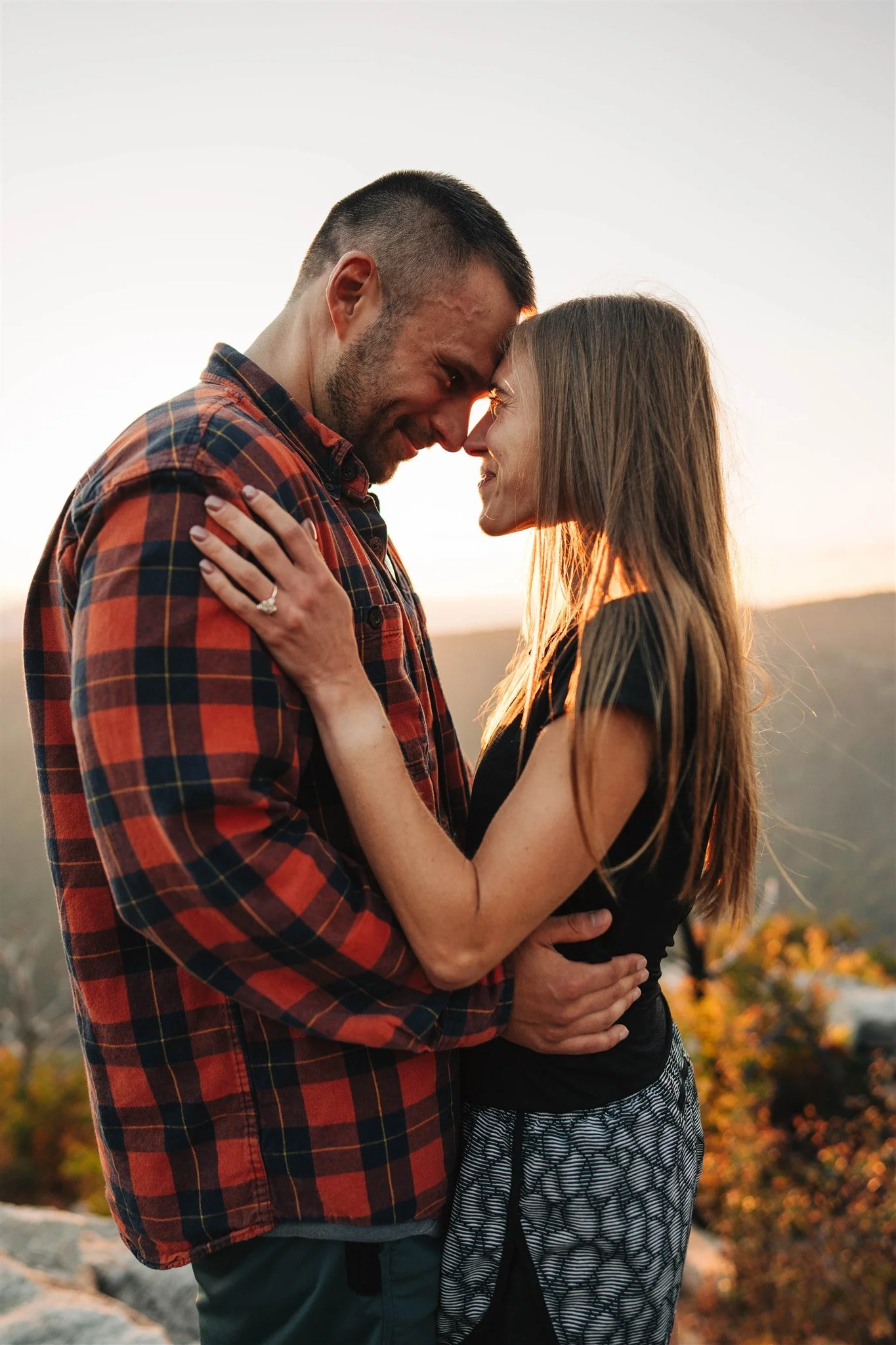 A couple standing close with foreheads touching, smiling at each other during sunset with a blurred landscape in the background.
