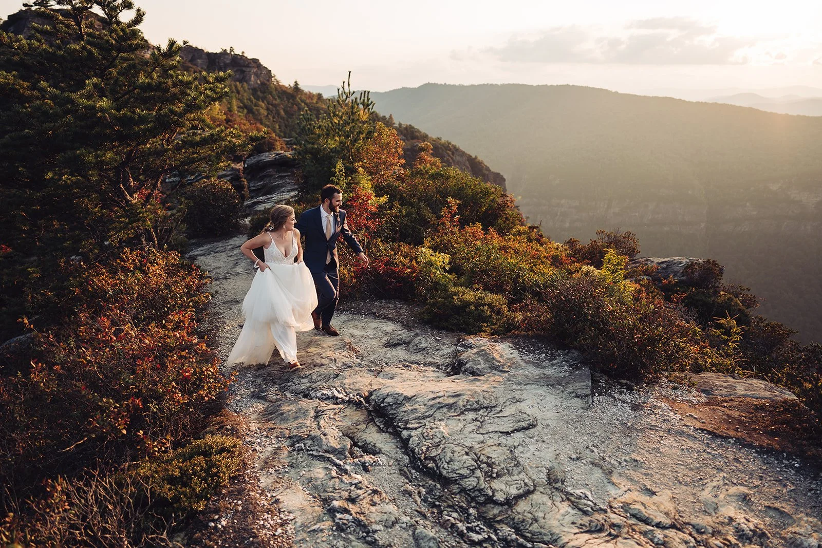 A bride and groom walking along a rocky mountain trail at sunset, surrounded by colorful autumn foliage with a mountain range in the distance.