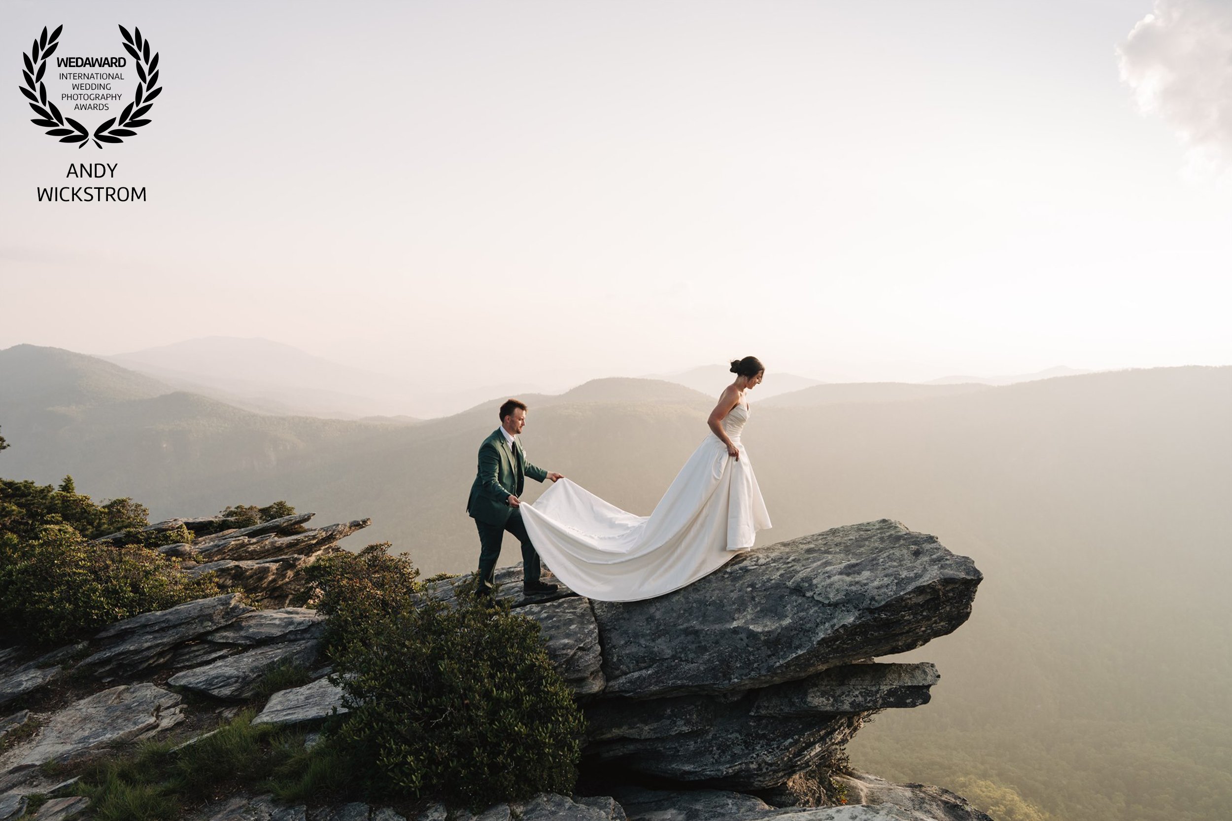 Bride walking up rock with train dress train being lifted by man in green suit at sunset.