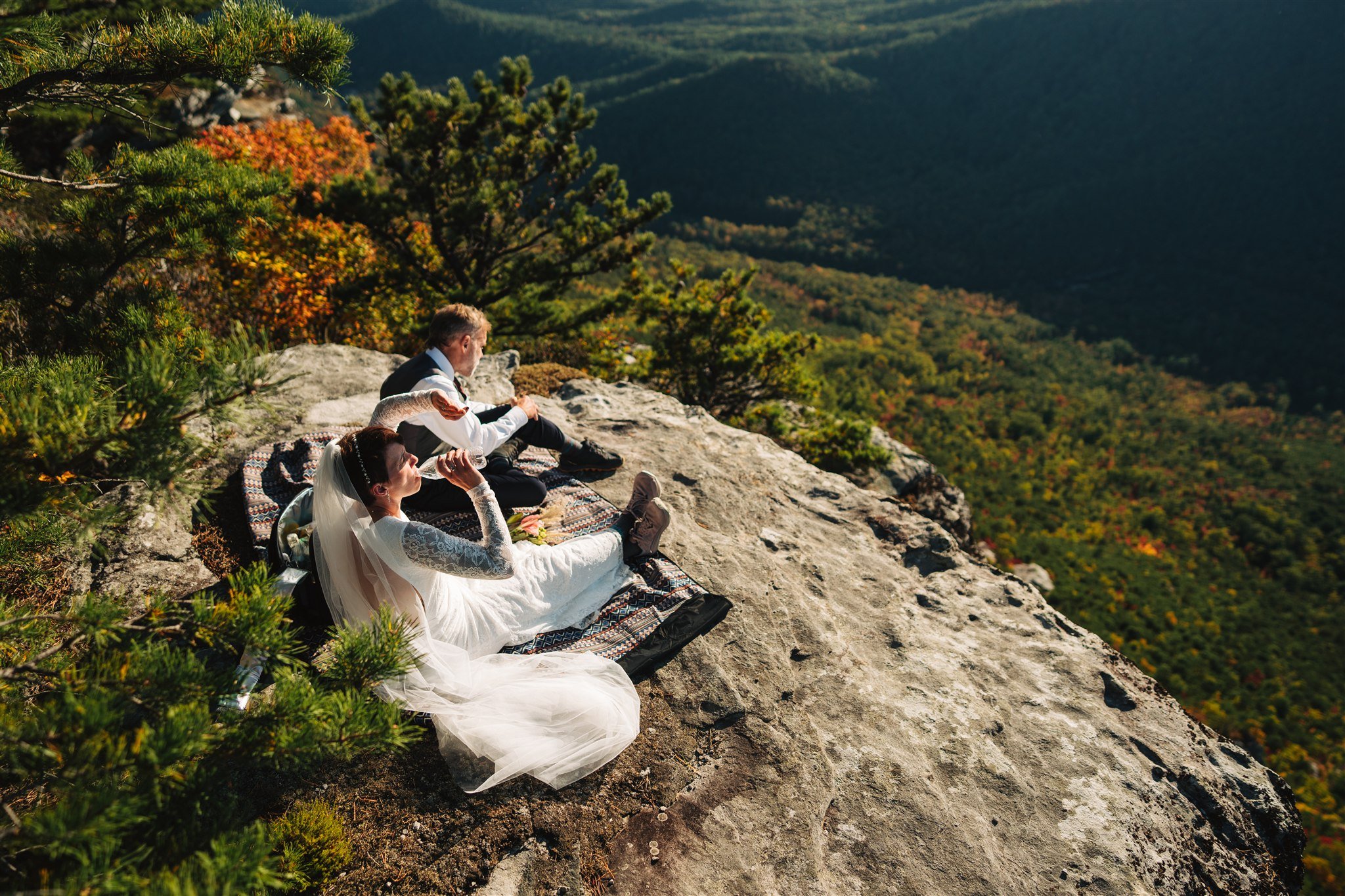 A bride and groom sitting on a rocky ledge overlooking a forested mountain landscape with autumn colors, during their wedding celebration.