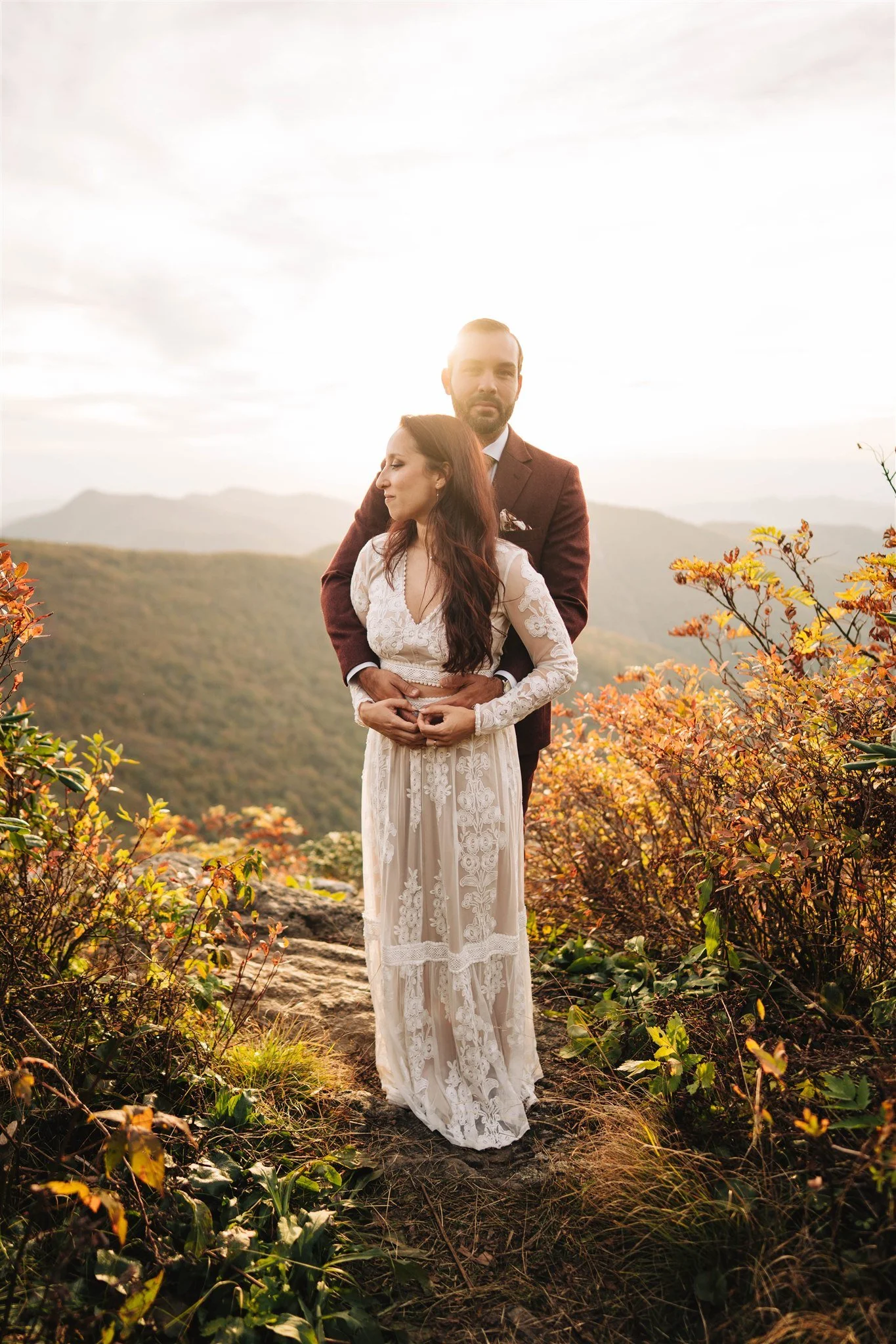 A couple dressed in formal wedding attire standing outdoors during sunset, with mountains and autumn foliage in the background.