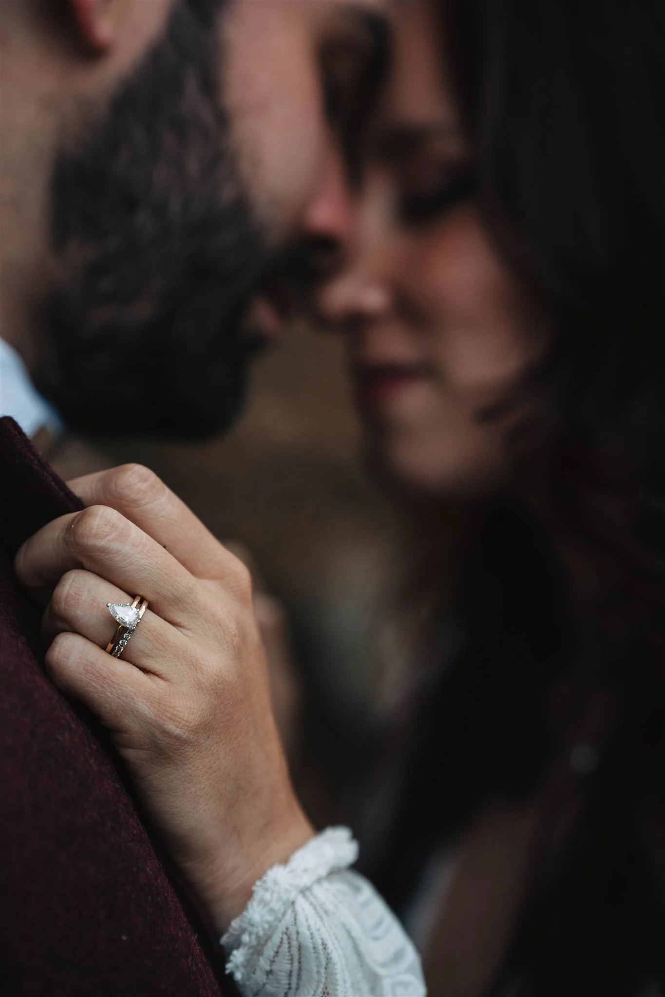 Close-up of a couple with their faces touching, woman holding the man's shoulder to show her engagement ring. The background is blurred.