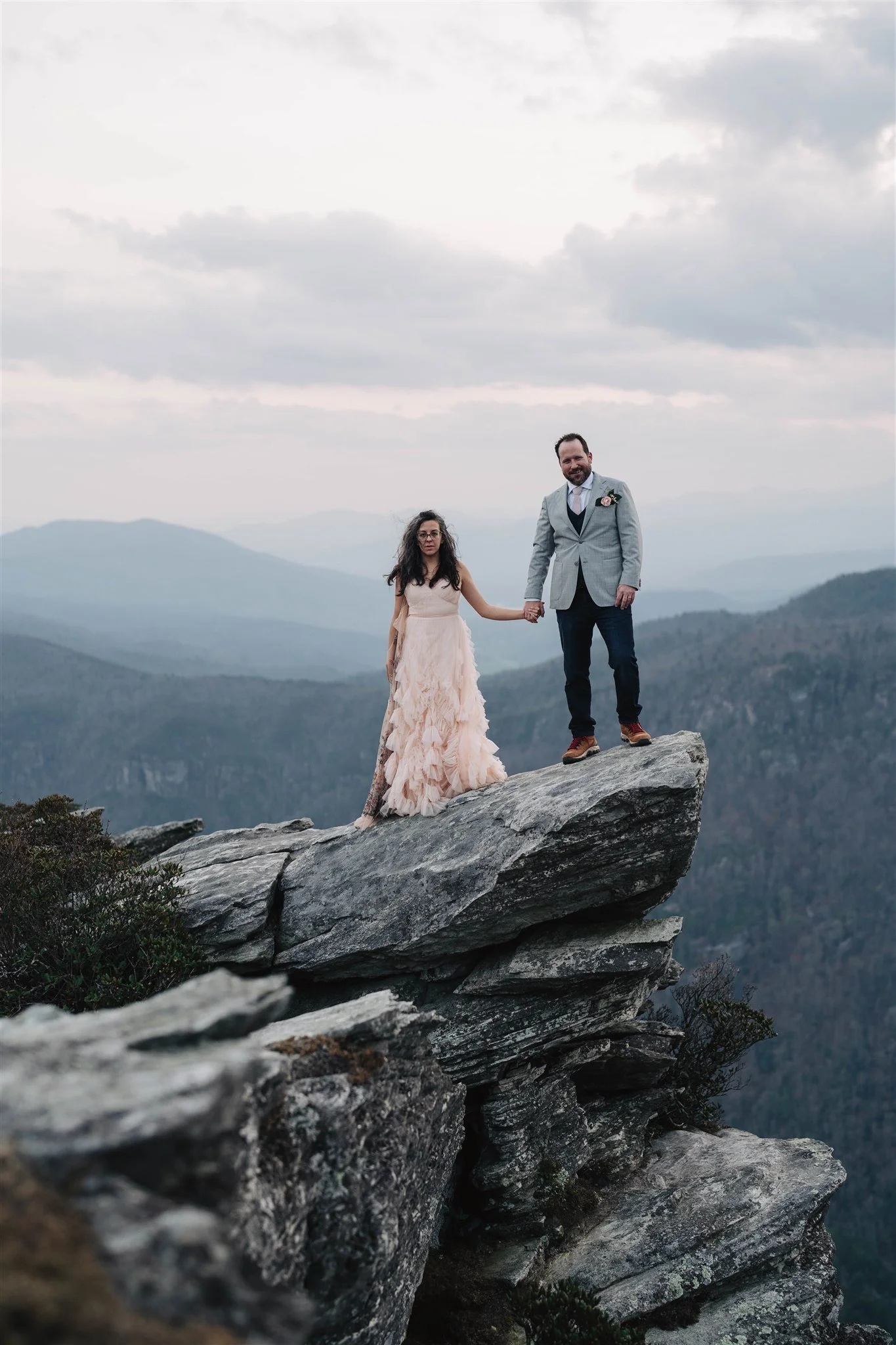 A bride and groom standing on a large rock formation in a mountainous landscape, holding hands, with misty mountains in the background.