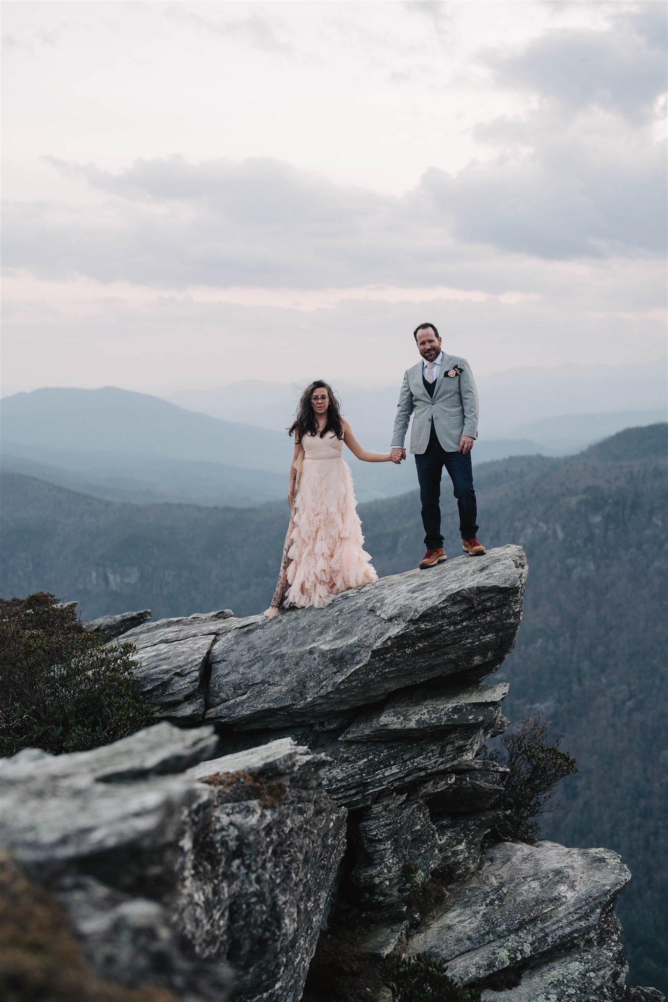 A couple standing on the edge of a rocky cliff, holding hands. The woman wears a flowing, light-colored dress, and the man is in a gray suit. The background features misty mountains under a cloudy sky.