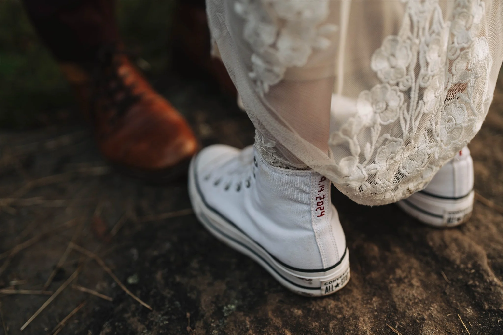 Close-up of a person's foot wearing a white Converse sneaker with black accents next to a boot, on dirt ground. The person is wearing a beige lace skirt.