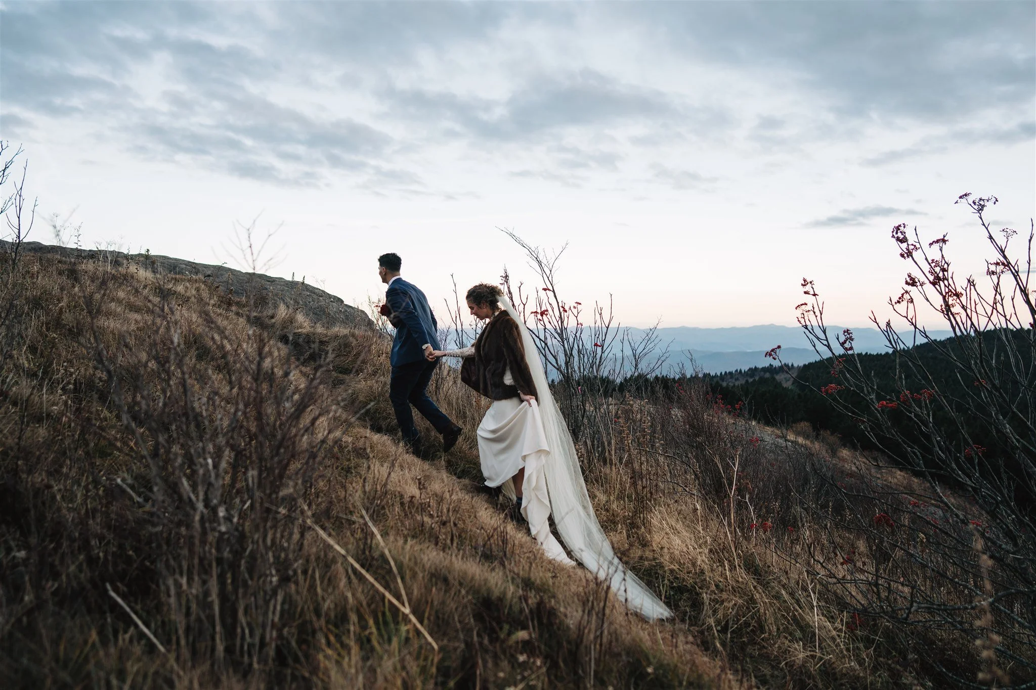 A bride and groom walking up a hillside trail in a mountainous landscape, holding hands, during sunset or late afternoon.