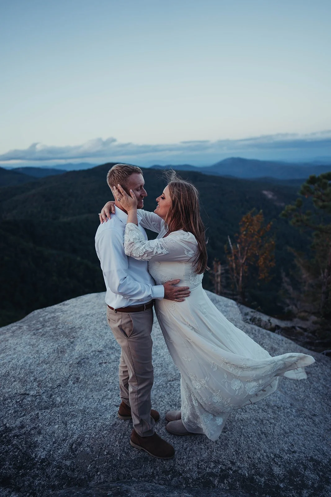 A couple standing on a large rock atop a mountain, embracing each other during sunset with a scenic vista of rolling hills and blue sky in the background.