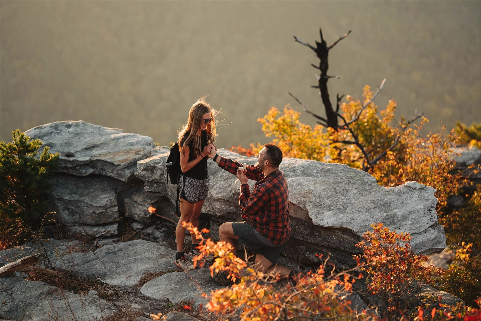 A man proposing marriage to a woman on a rocky outdoor mountain with fall foliage in the background.