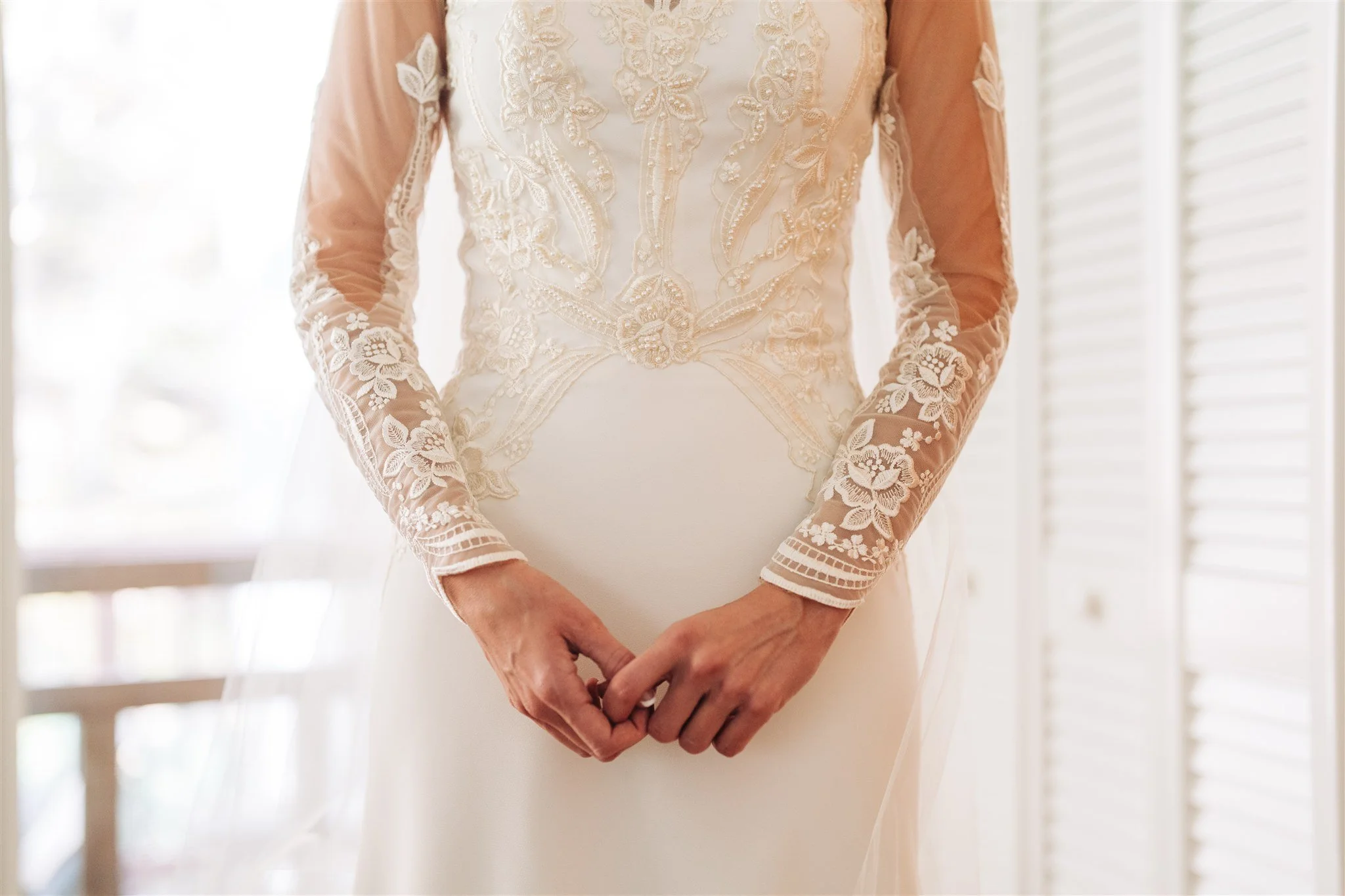 Close-up of a woman wearing a wedding dress with intricate lace embroidery on the bodice and sheer long sleeves, standing in front of a bright window with white blinds.