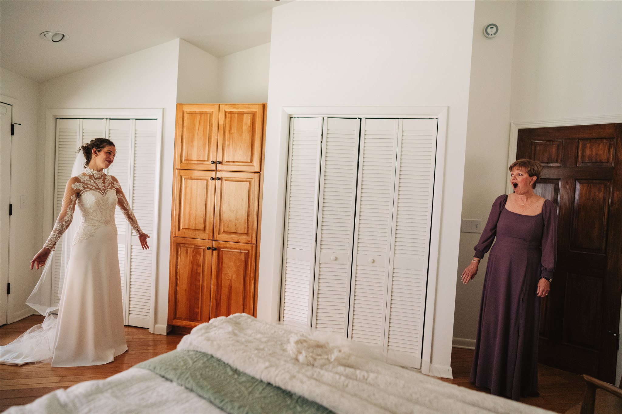A bride in a wedding dress smiling and gesturing with her arms, facing an older woman in a purple dress with an excited expression, inside a bedroom with white walls and wooden furniture.