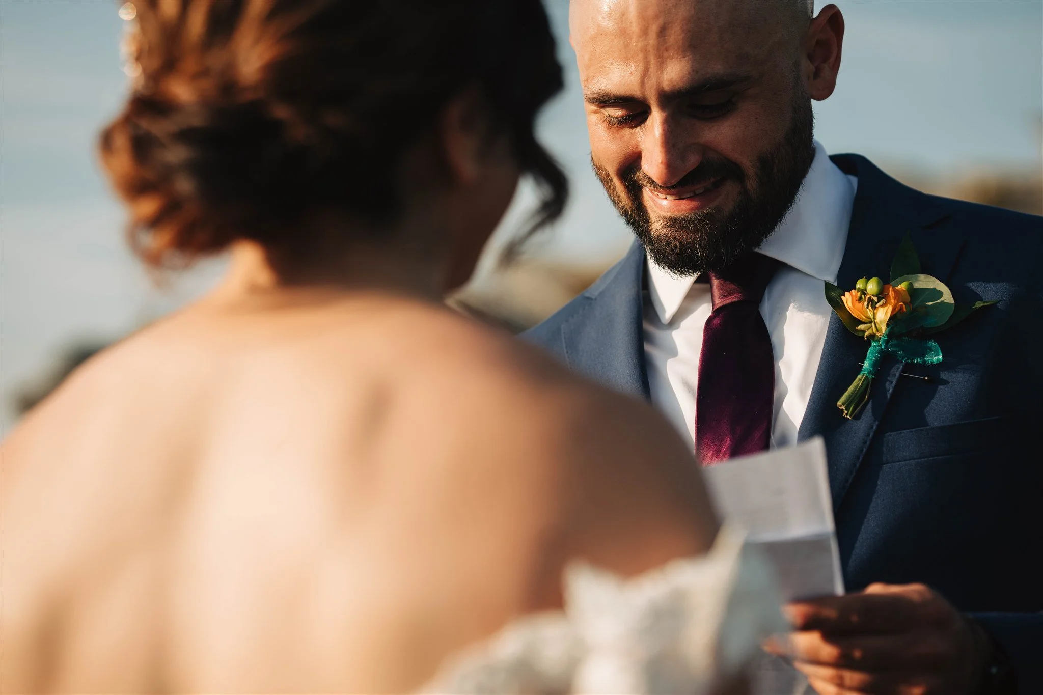 A man in a navy suit with a boutonniere is smiling at a woman during a wedding ceremony outdoors.