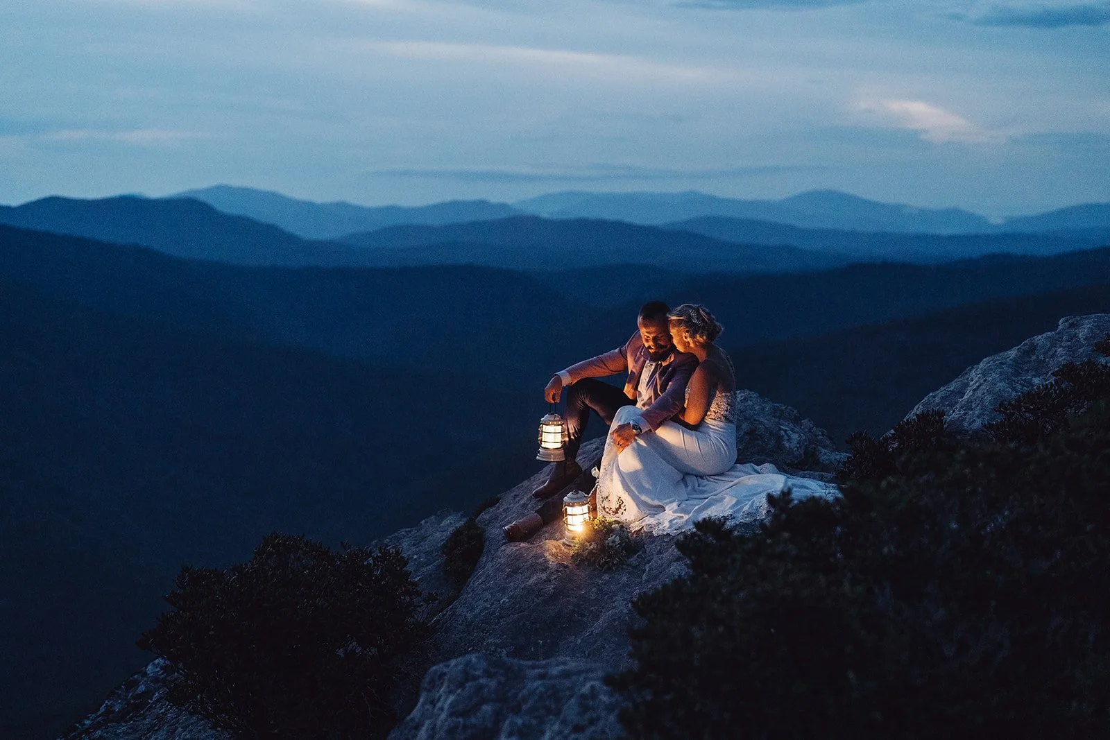 A couple dressed in wedding attire sitting on a rocky mountain edge at dusk, holding lanterns with a mountain range in the background.
