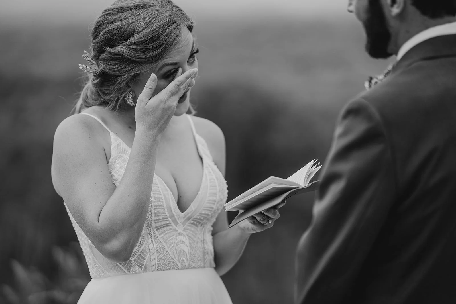 A woman in a wedding dress wiping away tears while reading vows from a book during her wedding ceremony, with a man in a suit standing in front of her.