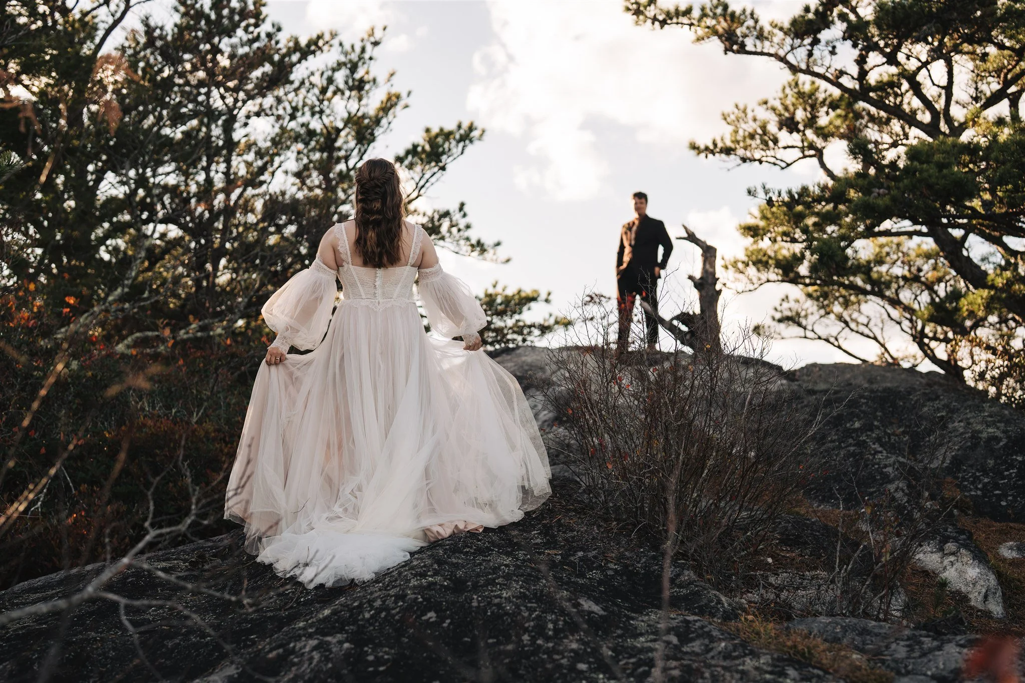 Woman in a white wedding dress walking up a rocky hill towards a man standing on the top, with trees and sky in the background.