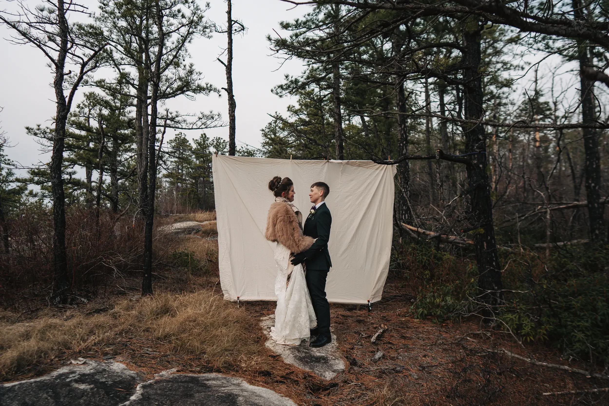 A couple in wedding attire holding hands and looking into each other's eyes in front of a white cloth backdrop in a wooded outdoor setting.