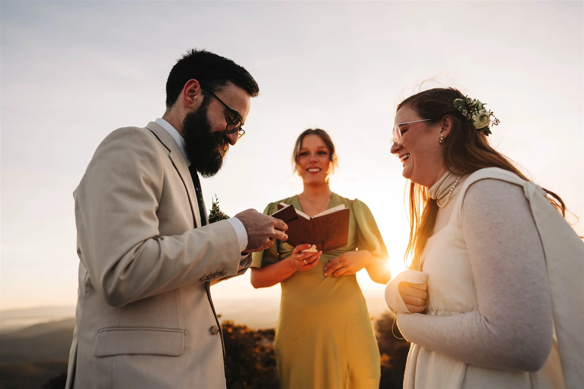A wedding ceremony outdoors at sunset with two people exchanging vows. A bearded man in a light suit is holding a notebook, while a woman in a white dress with flowers in her hair smiles. An officiant in a green dress stands between them holding a book. The sun sets in the background.