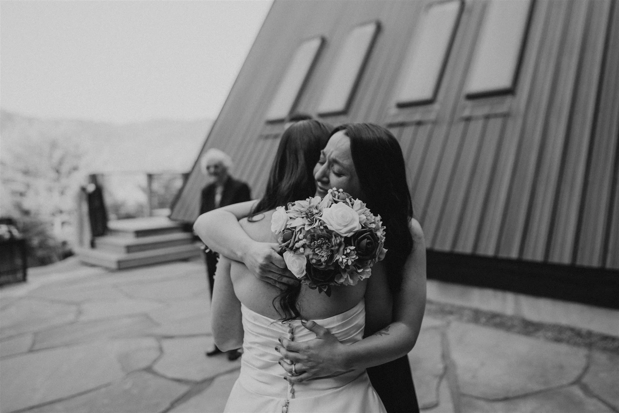 A woman in a wedding dress is hugging another woman, who is holding a bouquet of flowers, outside near a building.