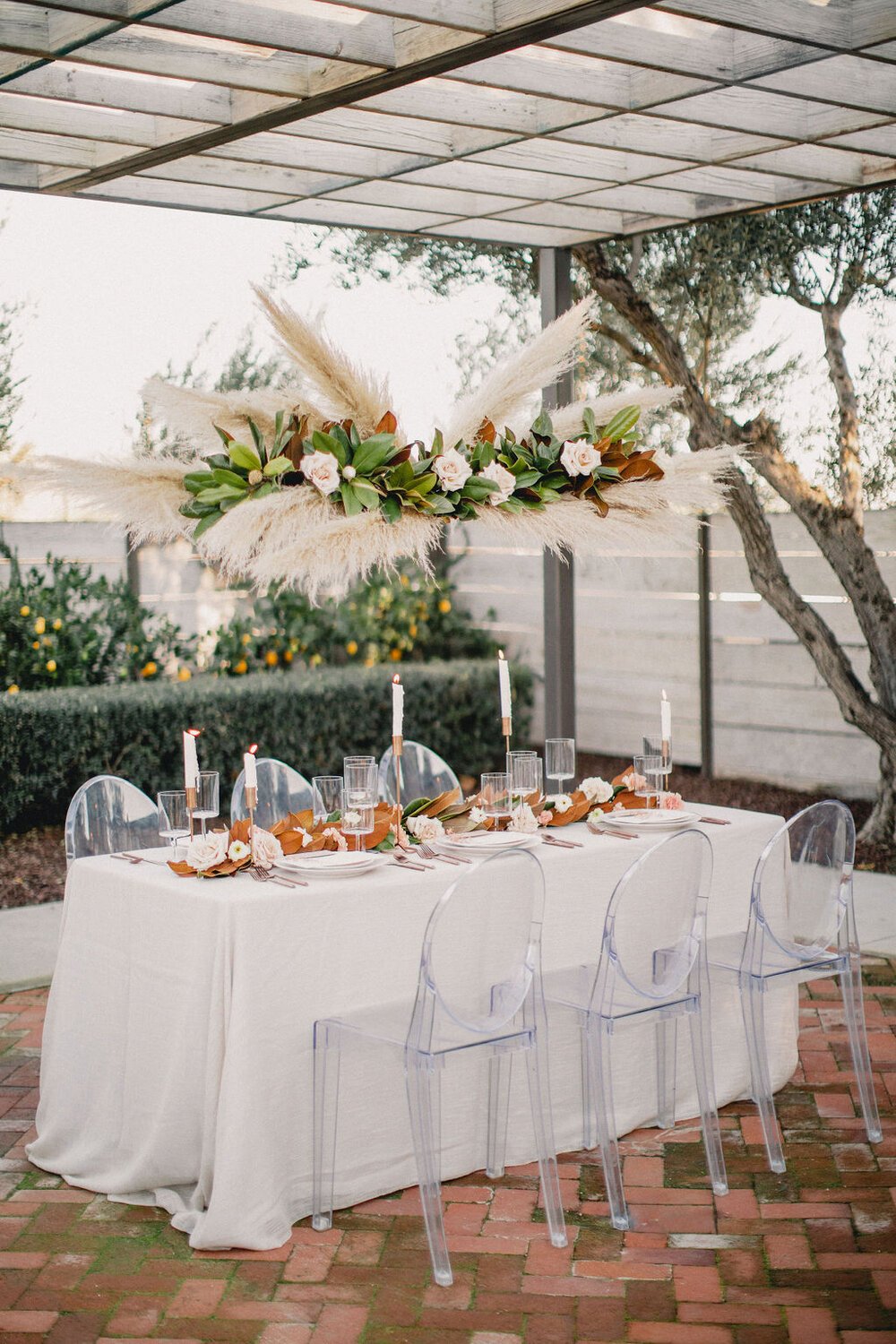 An outdoor dining table decorated with white tablecloth, floral centerpiece, candles, and glassware, set under a pergola with a large floral arrangement hanging above. Clear chairs are arranged around the table, with trees and a wooden fence in the background.
