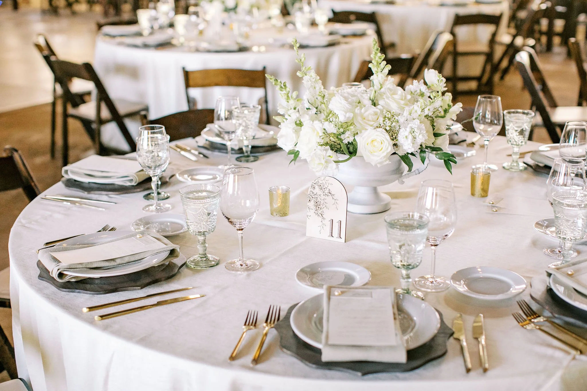 Elegant round table set with white tablecloth, floral centerpiece, glassware, plates, gold cutlery, white and black napkins, a table number card, and small candles, at a formal event or wedding reception.