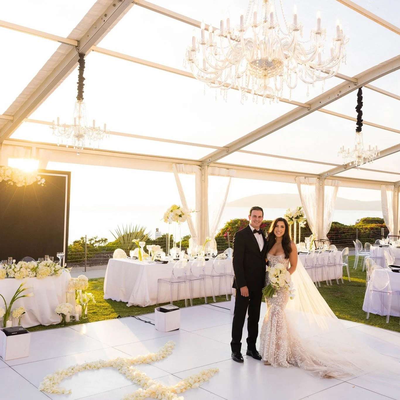 A bride and groom standing together at their wedding reception in a white tent with chandeliers. The bride is holding a bouquet of white flowers, and the groom is wearing a black tuxedo. The reception area is decorated with white flowers and candles, with the sunset visible in the background.