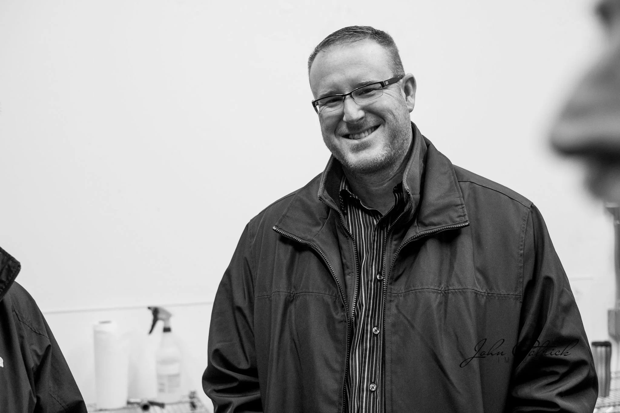 A smiling man with glasses, short hair, and a beard, wearing a dark jacket over a collared shirt, standing indoors against a plain wall.